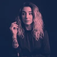 A young woman with long, wavy blonde hair, wearing a black top, posing against a dark background.