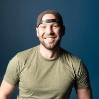 Men smiling with a beard, wearing a backwards cap and casual green t-shirt, against a blue background.