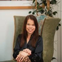 Woman with long brown hair smiling, sitting in a high-back green chair in a room with houseplants and framed art on the wall.