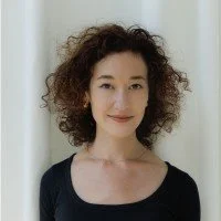 A woman with curly hair wearing a black top standing in front of a plain background.