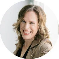 Woman with wavy light brown hair smiling, wearing a brown jacket, in a studio with a white background.