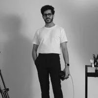 A man wearing glasses, a white t-shirt, and black pants, standing in a room with a table and some objects on it.
