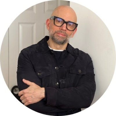A middle-aged man with a bald head and beard, wearing glasses and a black jacket, standing in front of a white door with his arms crossed.