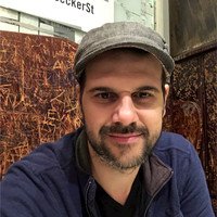 A man with a beard and mustache wearing a gray cap and navy blue jacket, sitting indoors near a brown wooden wall and a white sign.
