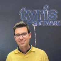 Young man with glasses wearing a yellow shirt standing in front of a wall with the Tyrnis Software logo.