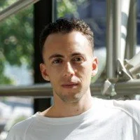 A man with short dark hair and light skin wearing a white shirt, sitting outdoors near a bus or large vehicle.