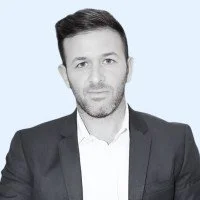 Portrait of a man with dark hair and beard wearing a suit and white shirt against a light blue background.