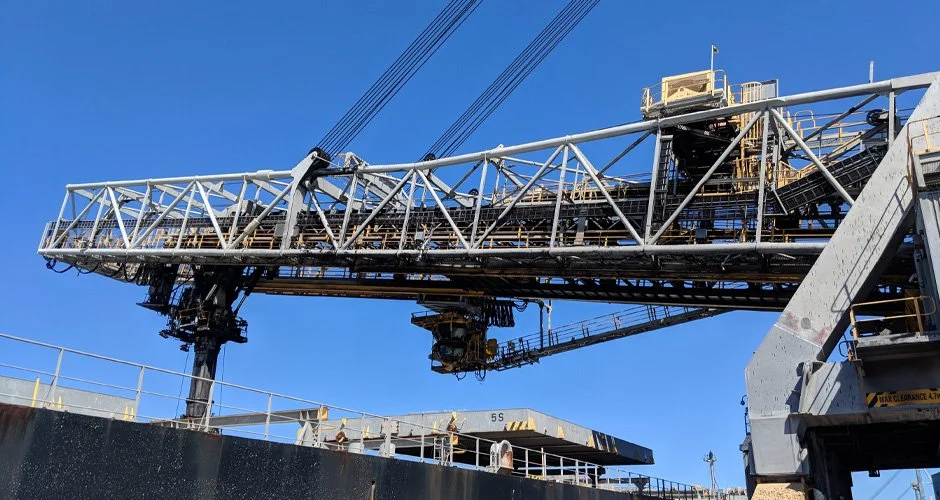 Close-up of an industrial conveyor crane structure against a clear blue sky, with safety railings and machinery below.