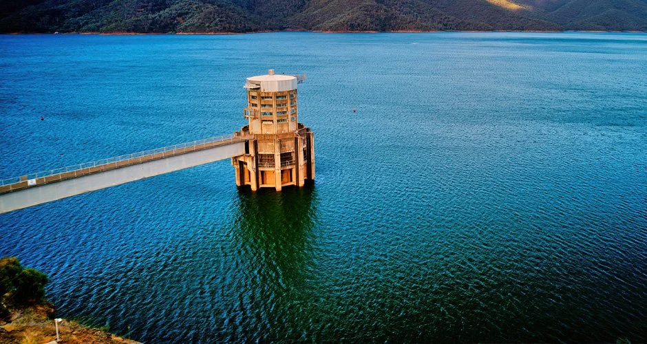 A dam structure extending into a large body of water, with surrounding green hills and mountains in the background.