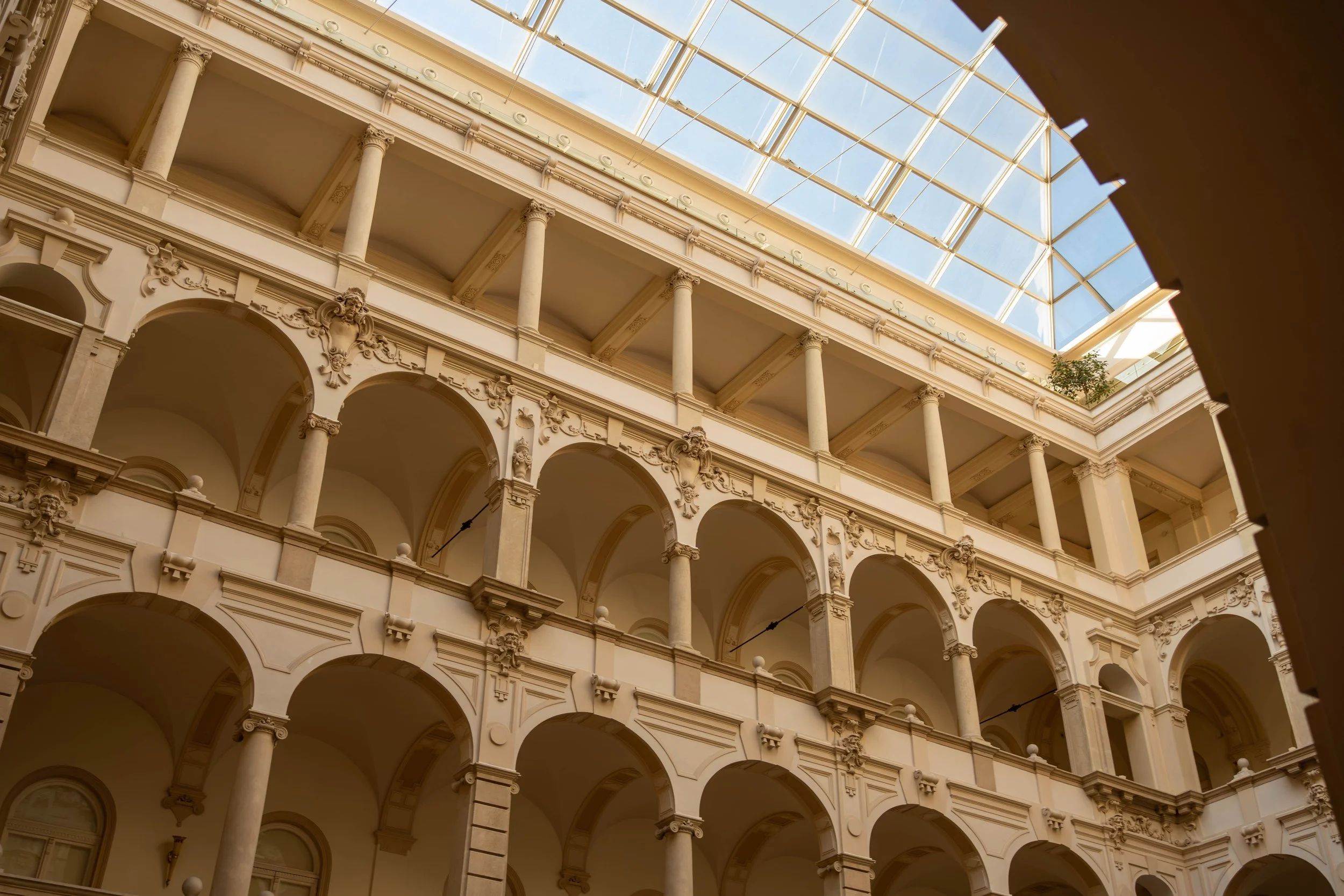 Three levels of beige, ornate architecture with arches, columns, and decorative elements inside a building, topped by a large glass skylight.