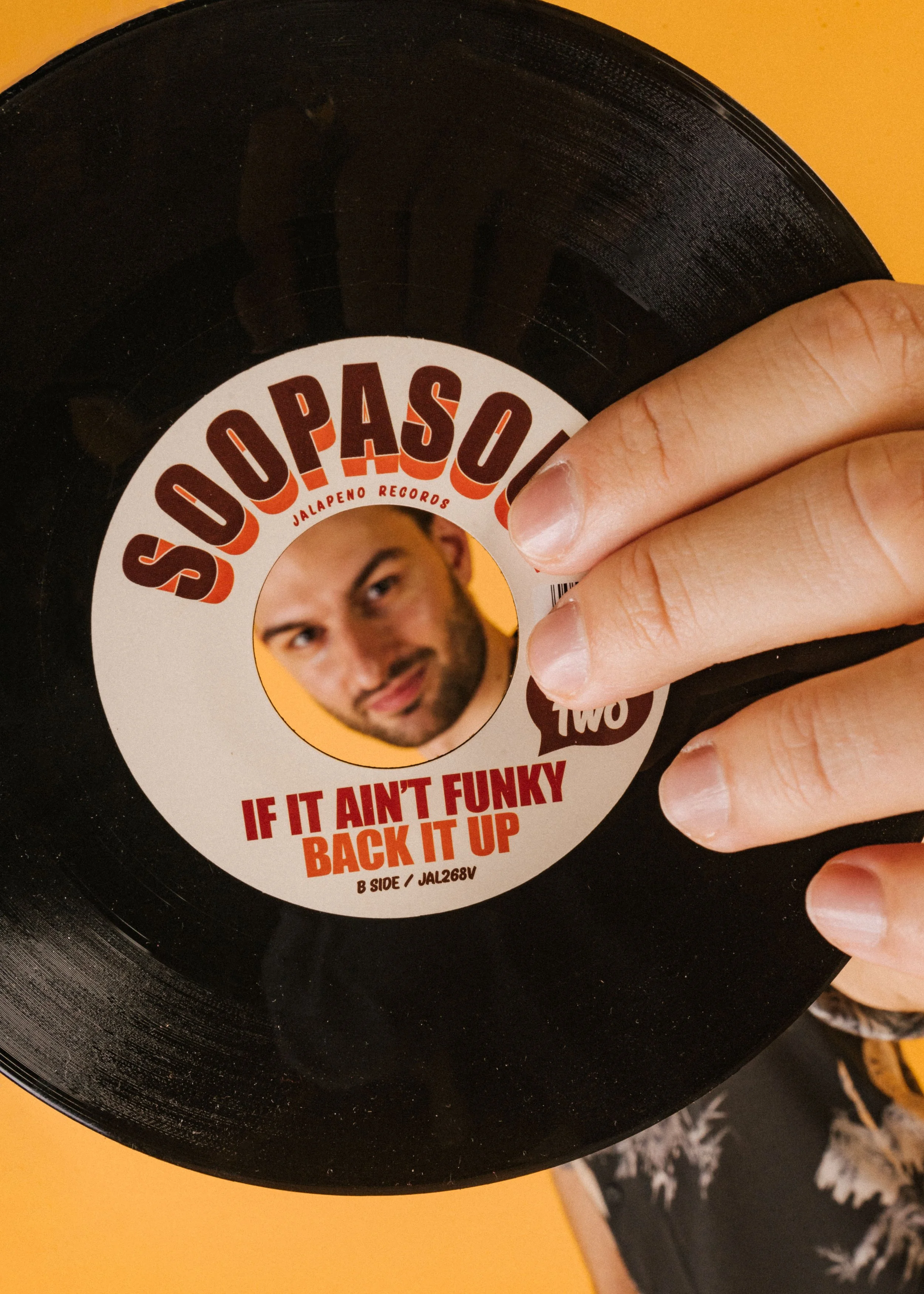 Close-up of a vinyl record with a label featuring a man's face and the text 'Souo Pasol' and 'Jalapeno Records'. The record is held by a person's hand with the thumb and index finger visible, against an orange background.