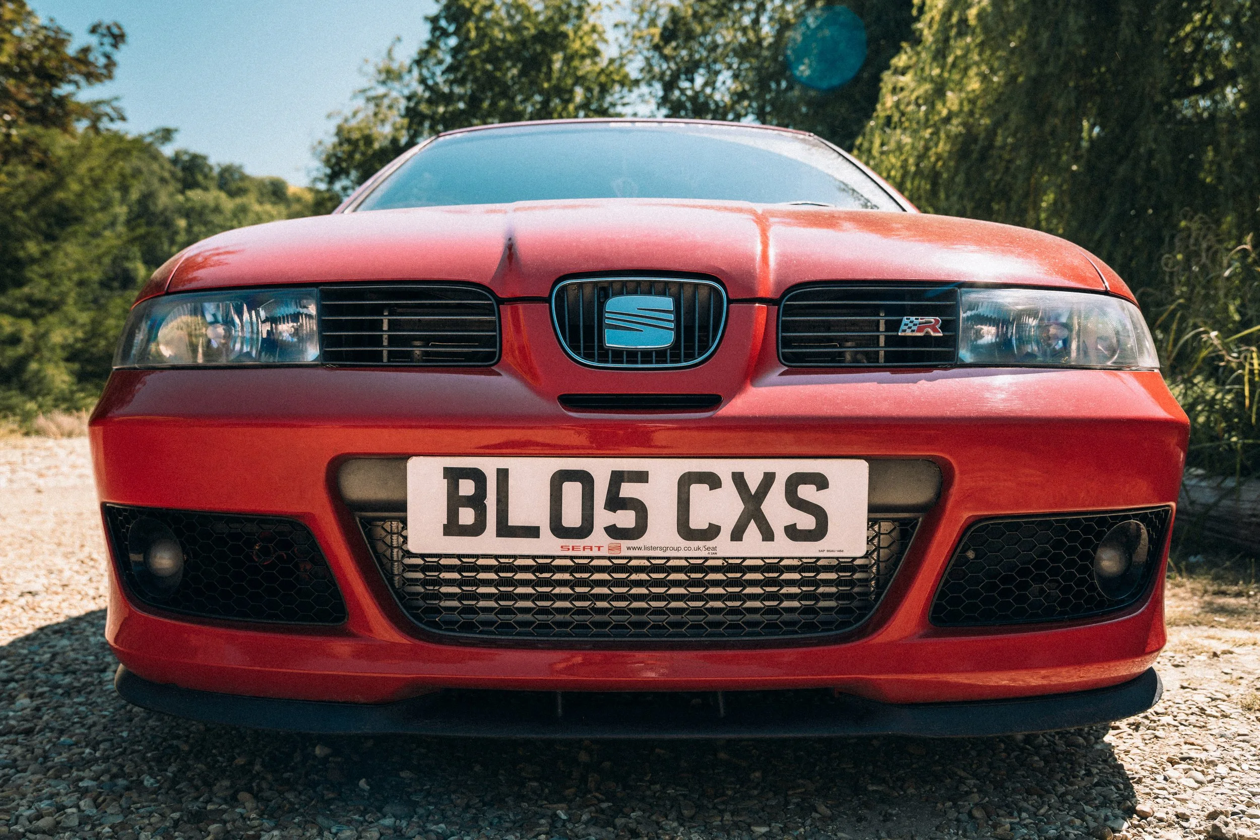 Red sports car with a black grille and license plate reading 'BL05 CXS' parked on a gravel surface, surrounded by green trees and blue sky.