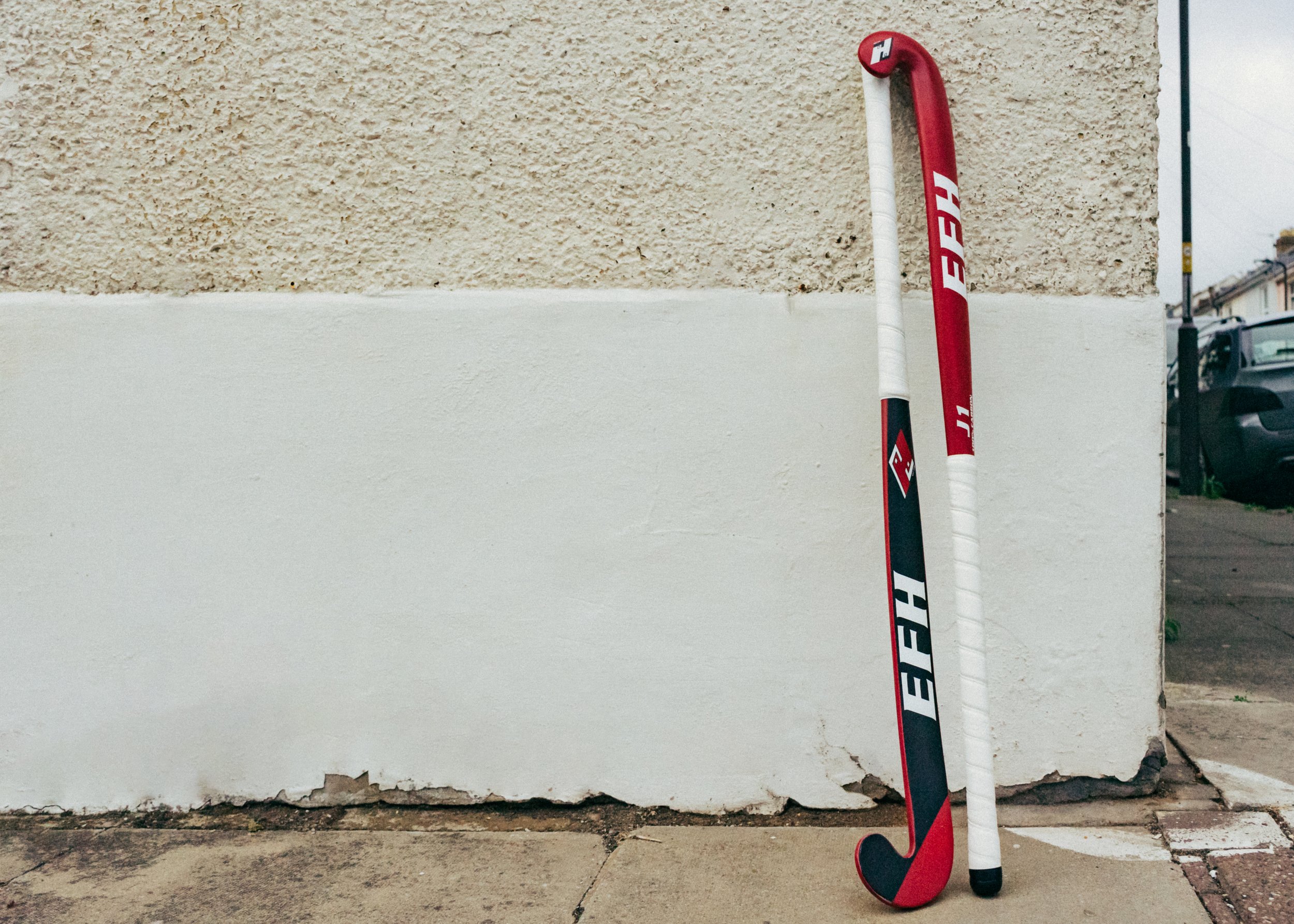 A hockey stick leaning against a white wall near a beige textured wall, with a parking lot and cars visible in the background.