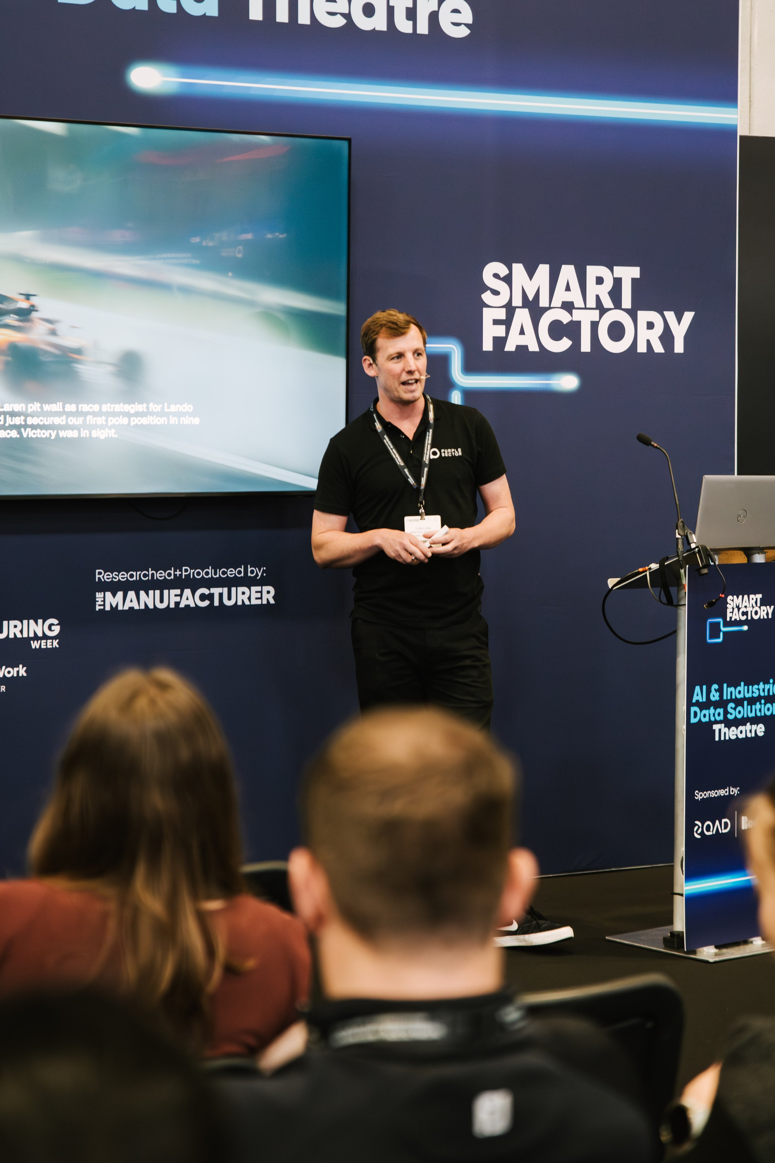 A man is giving a presentation at the Smart Factory event, standing before a large screen displaying a roller coaster race with text about a race strategist. Audience members are visible in the foreground, sitting and listening.