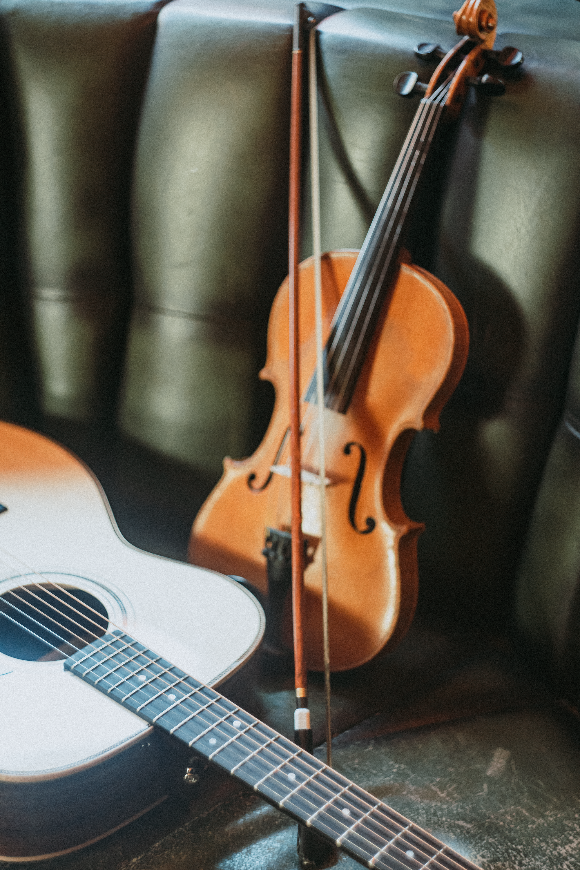 A close-up of a black acoustic guitar in the foreground and a wooden violin resting against a black leather couch in the background.