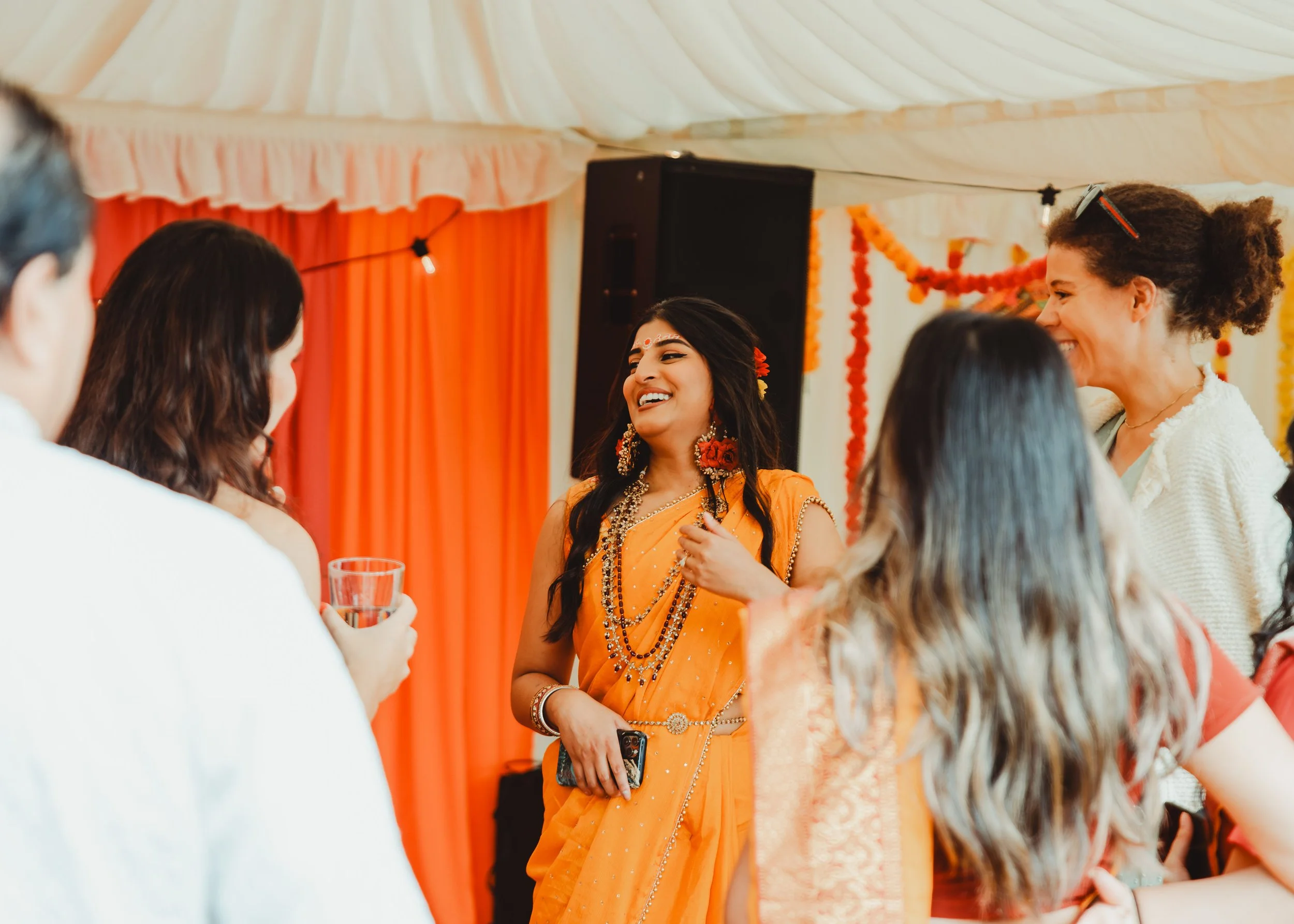 A group of women gathered at a celebration, with one woman dressed in traditional Indian attire smiling and holding a phone, others are laughing and talking, in a decorated indoor setting with orange and red drapes.