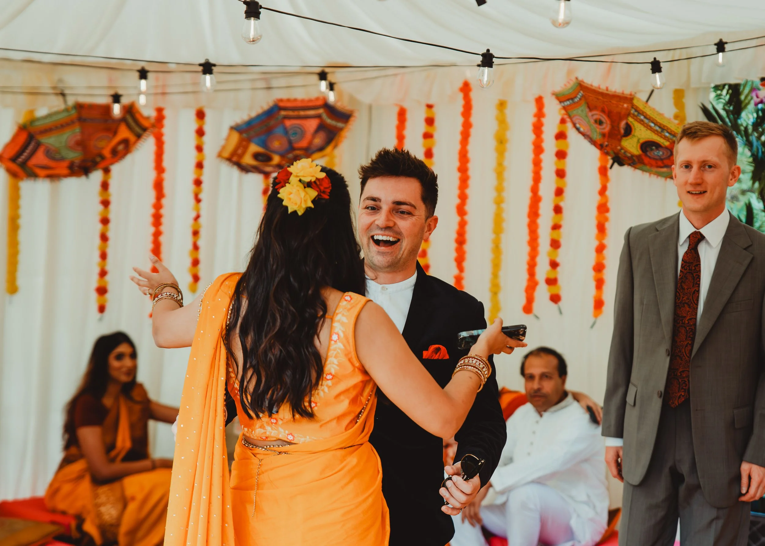 People celebrating at a colorful decorated event, with bright orange and yellow marigold flower garlands, traditional umbrellas, and string lights hanging from the ceiling.