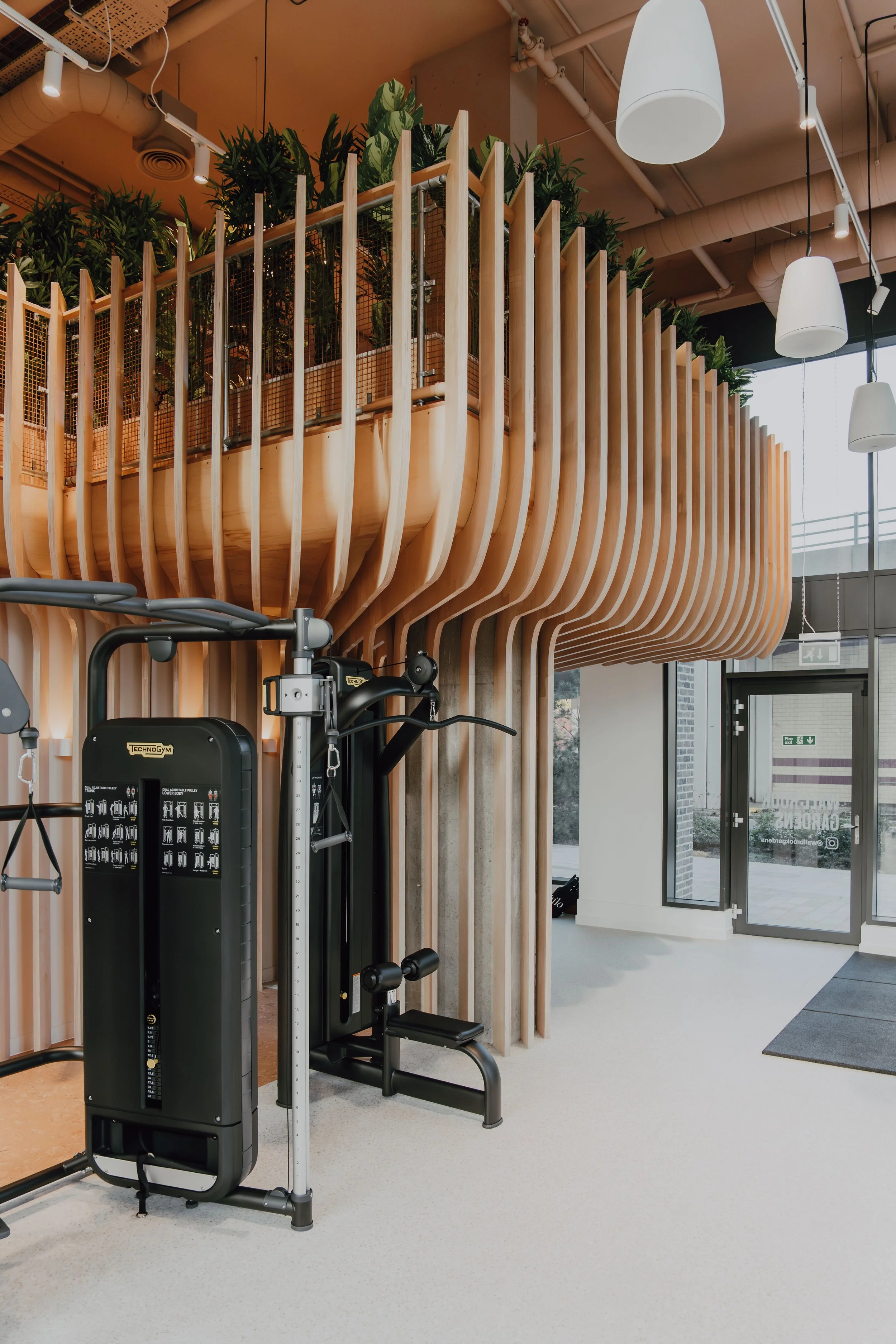 Gym area with fitness equipment in front of a wooden structure with plants on top, large windows, and ceiling-mounted lights