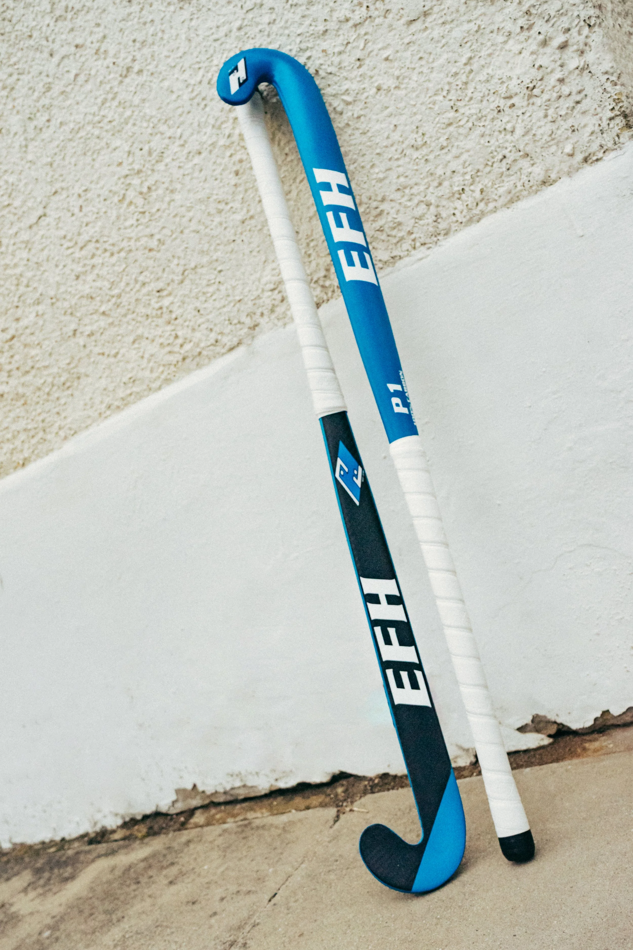 A blue and black hockey stick leaning against a white wall with a textured surface.