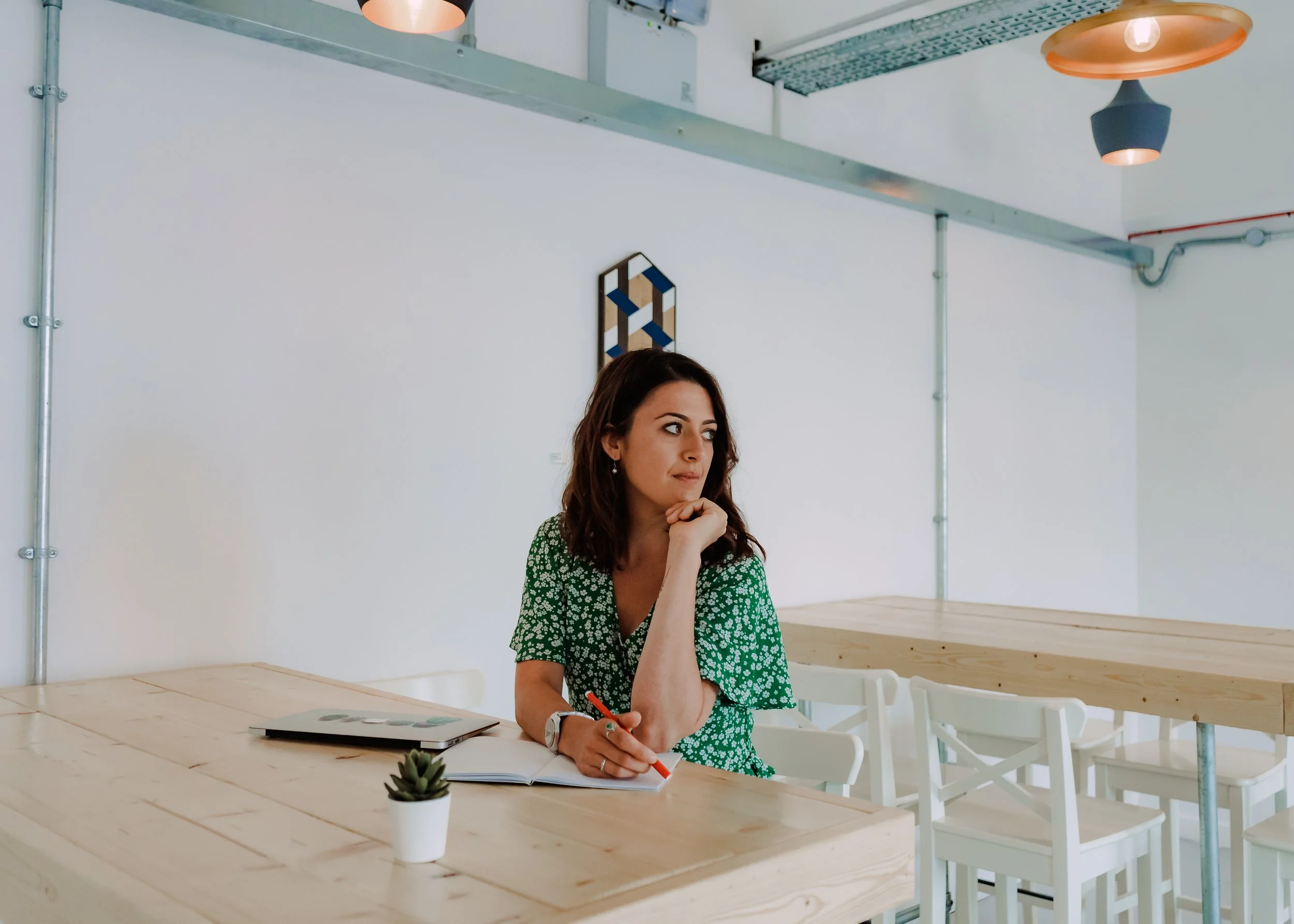 A woman with dark brown hair in a green floral dress sitting at a wooden table in a modern, minimalist room with white walls and white chairs. She has a contemplative expression, with her chin resting on her hand, and is holding a red pen over an open notebook. There is a small potted succulent plant and a closed laptop on the table, and framed geometric wall art on the white wall behind her.