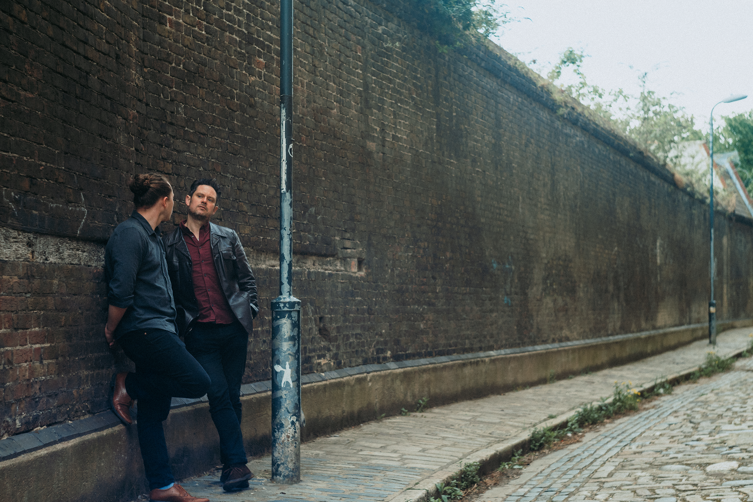 Two people, a man and a woman, leaning against a brick wall on a sidewalk. They are facing each other, having a conversation. The woman has her hair in a bun and is wearing a dark jacket. The man has dark hair, a beard, and is wearing a leather jacke