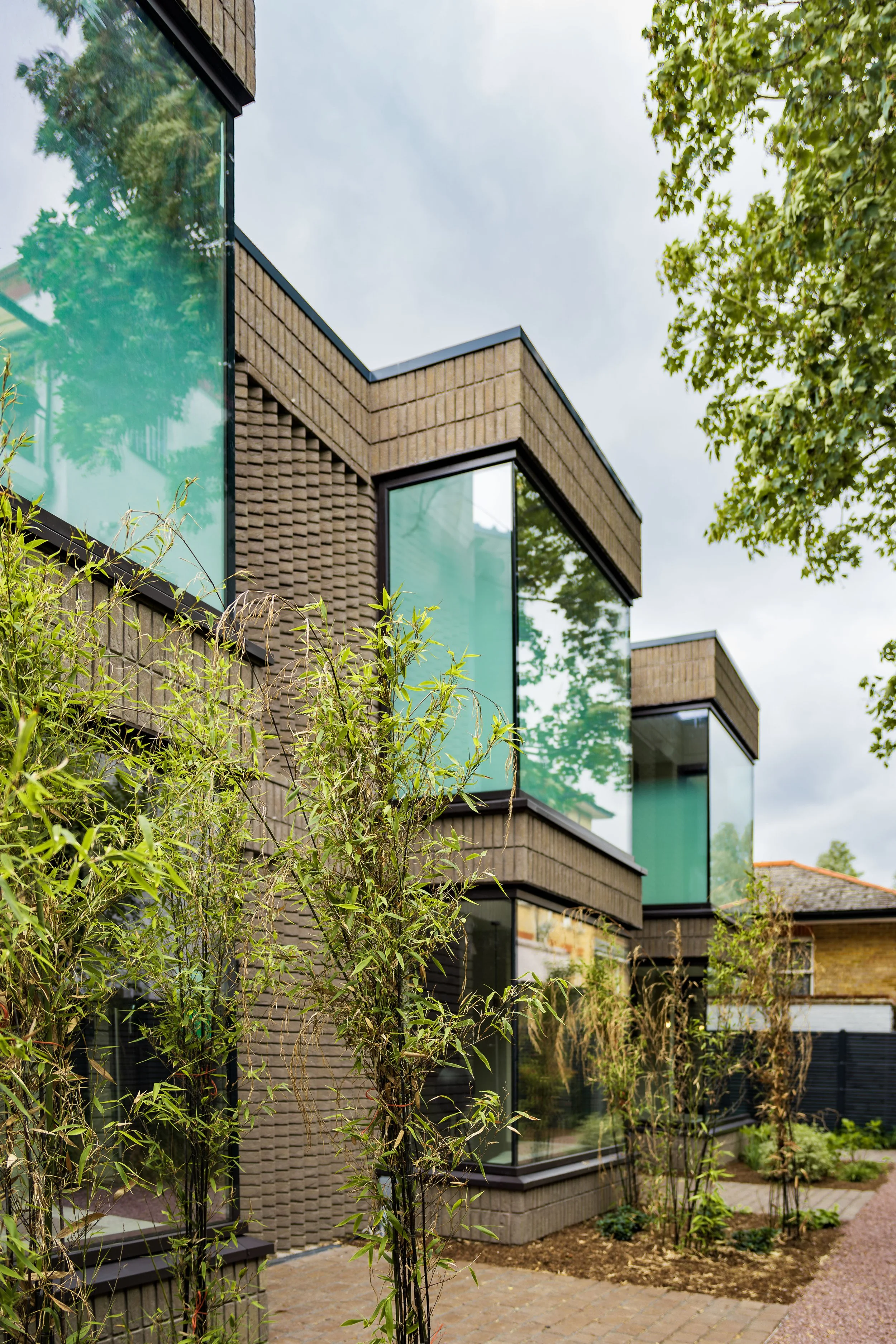 Modern residential building with large glass windows, brick facade, and greenery in the foreground, under a cloudy sky.