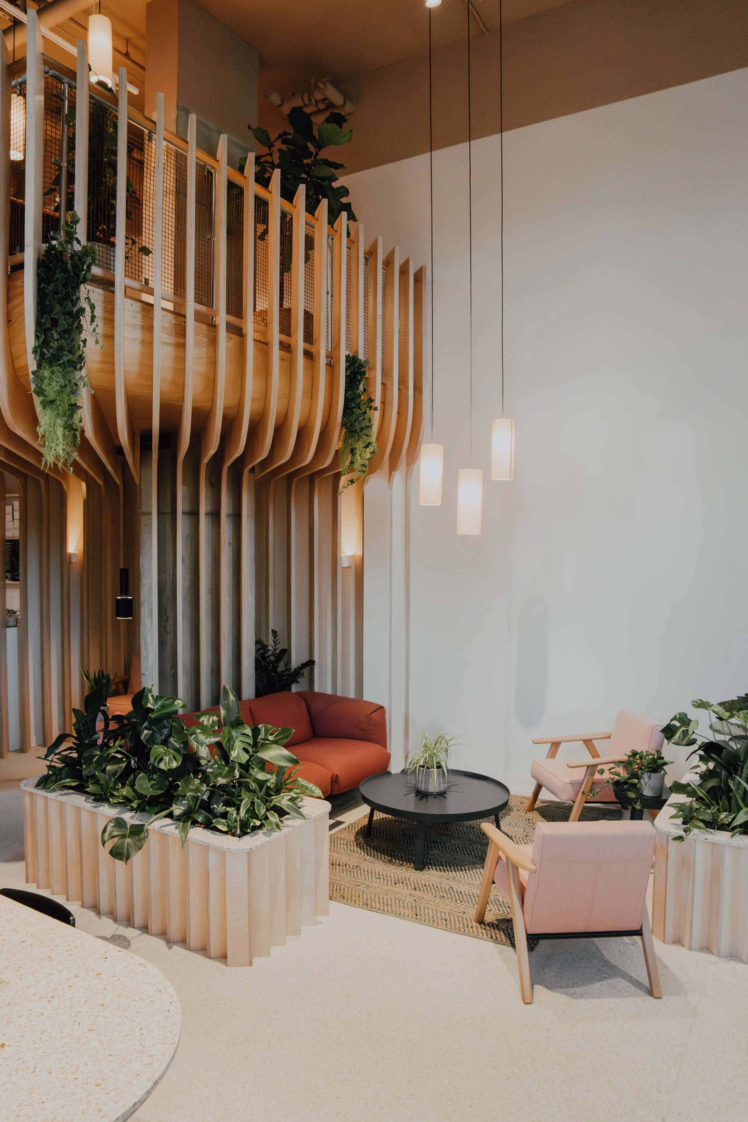 A modern indoor lounge area with pastel pink chairs, a black coffee table, a blush pink sofa, indoor plants, and hanging pendant lights, featuring wooden architectural accents and a light-colored floor.