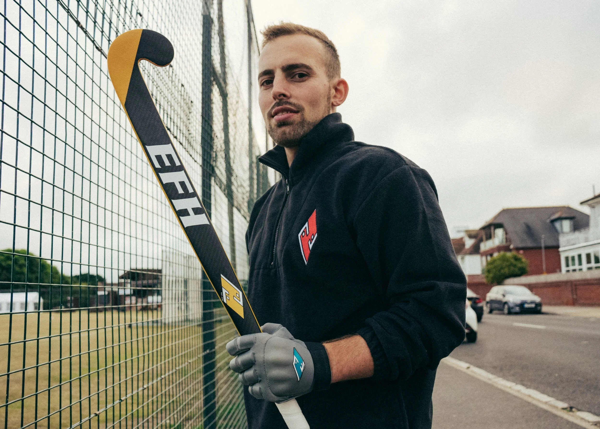 A man standing next to a metal fence on a sports field, holding a hockey stick, wearing gloves, and looking at the camera.