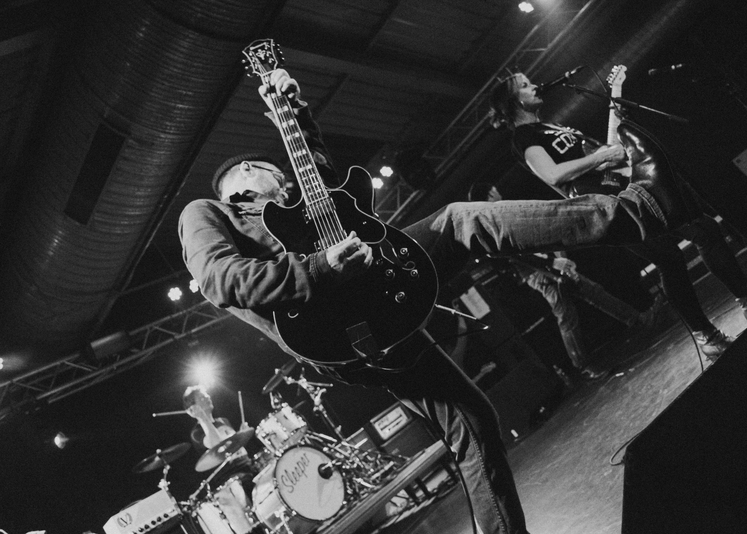 Black and white photo of a band performing on stage, featuring a guitarist, a drummer, and a singer with a microphone. The guitarist is in the foreground, playing an electric guitar, and the drummer is in the background behind a drum kit. The singer is seated with a microphone in front of them.