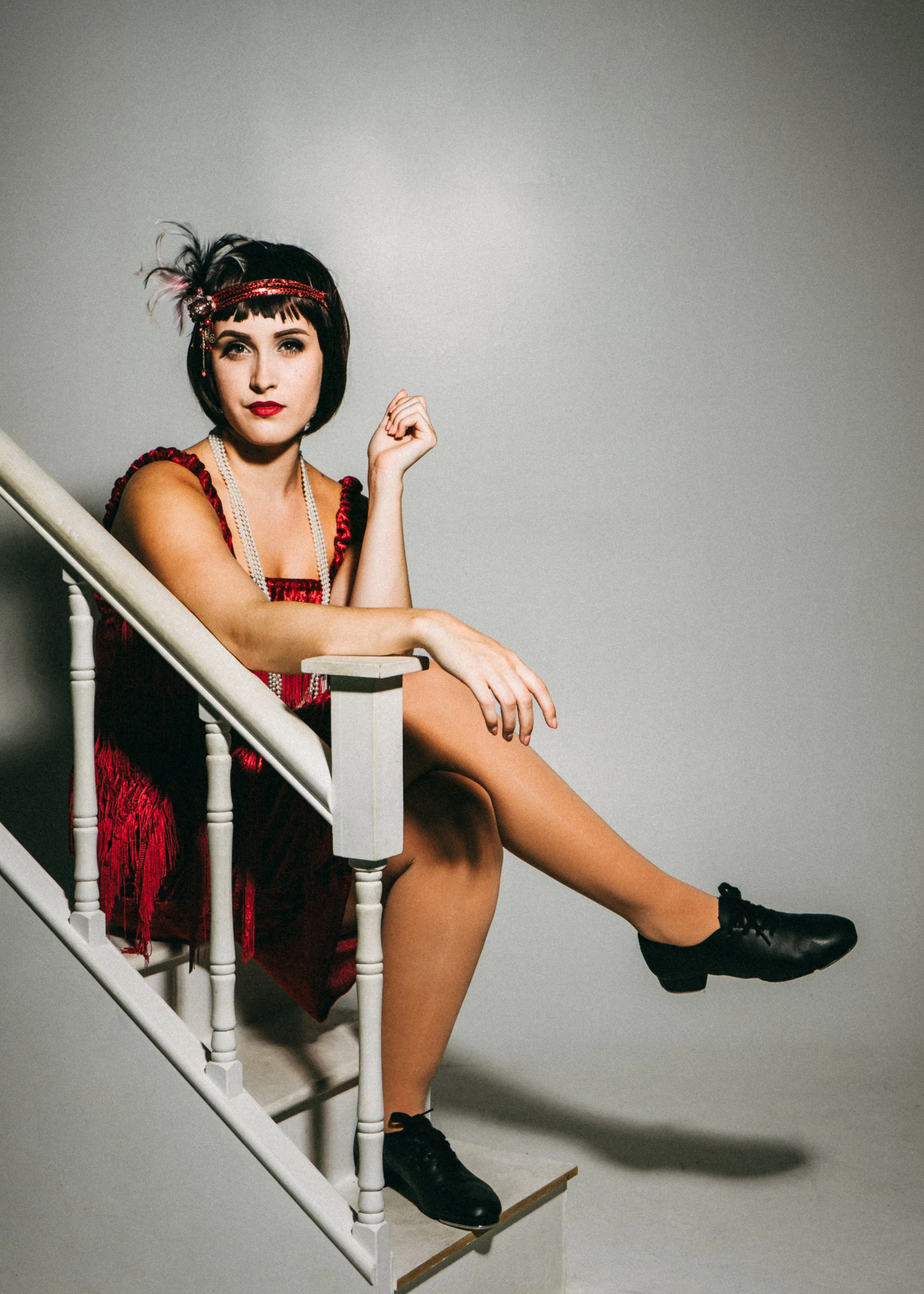 Woman with dark hair and fringe, wearing a red dress with fringes and accessories, sitting on a staircase with one leg crossed over the other, against a plain background.