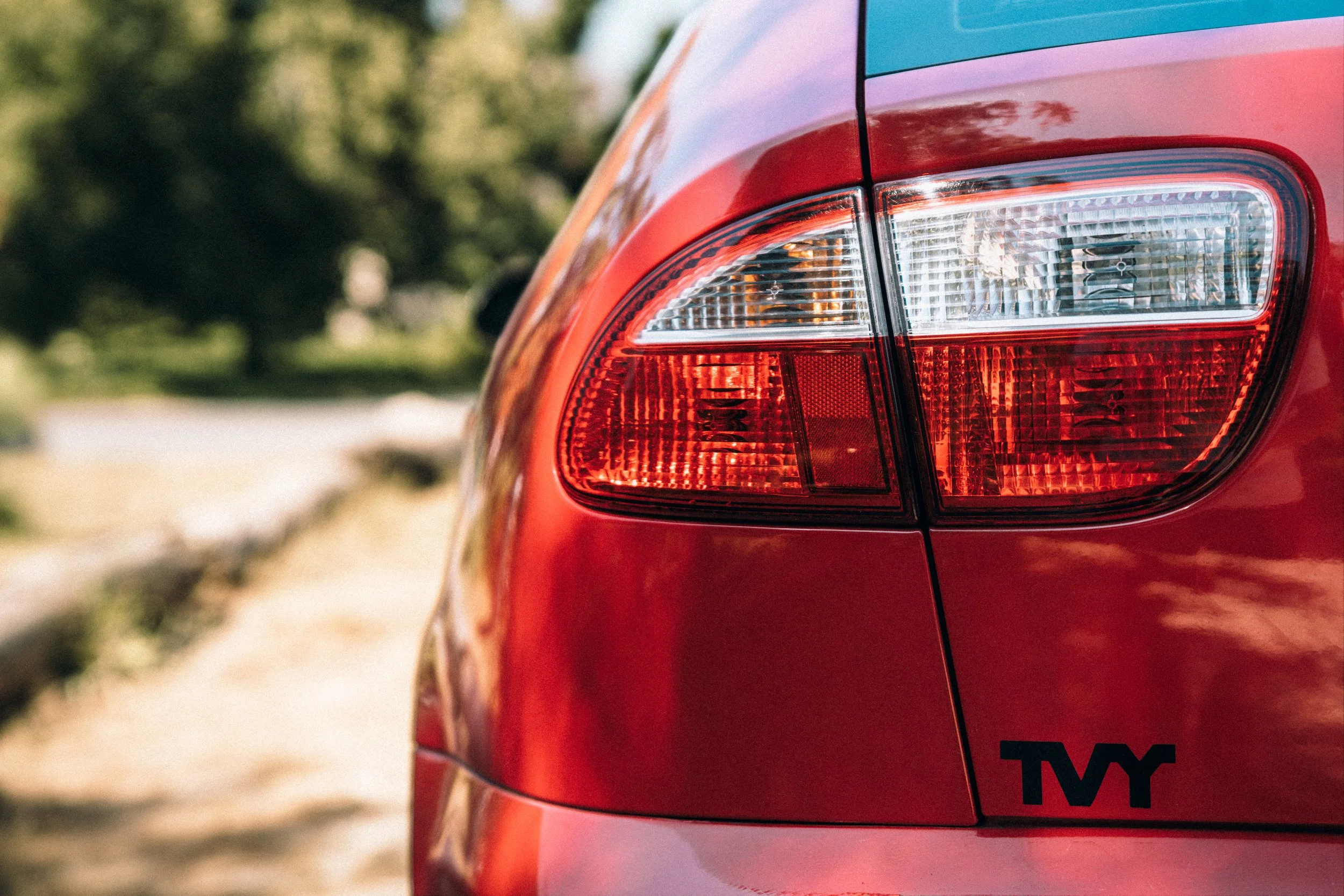 Close-up of the rear end of a red car showing the tail light and part of a sticker with black letters 'TVY'.