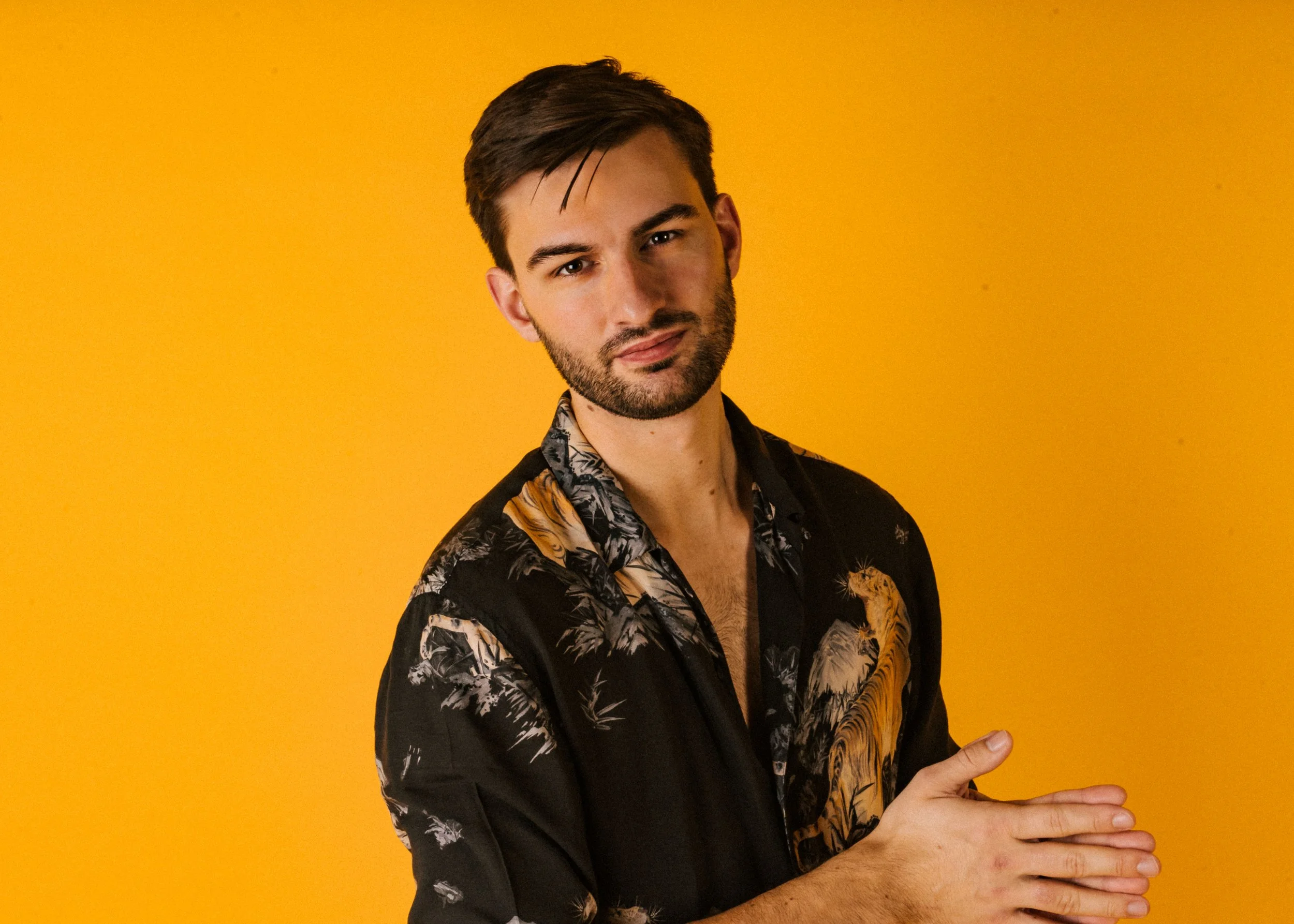 A young man with dark hair, a beard, and a mustache, wearing a black shirt with a tropical print, is standing against a yellow background. He has a neutral expression and is holding his hands together in front of his body.