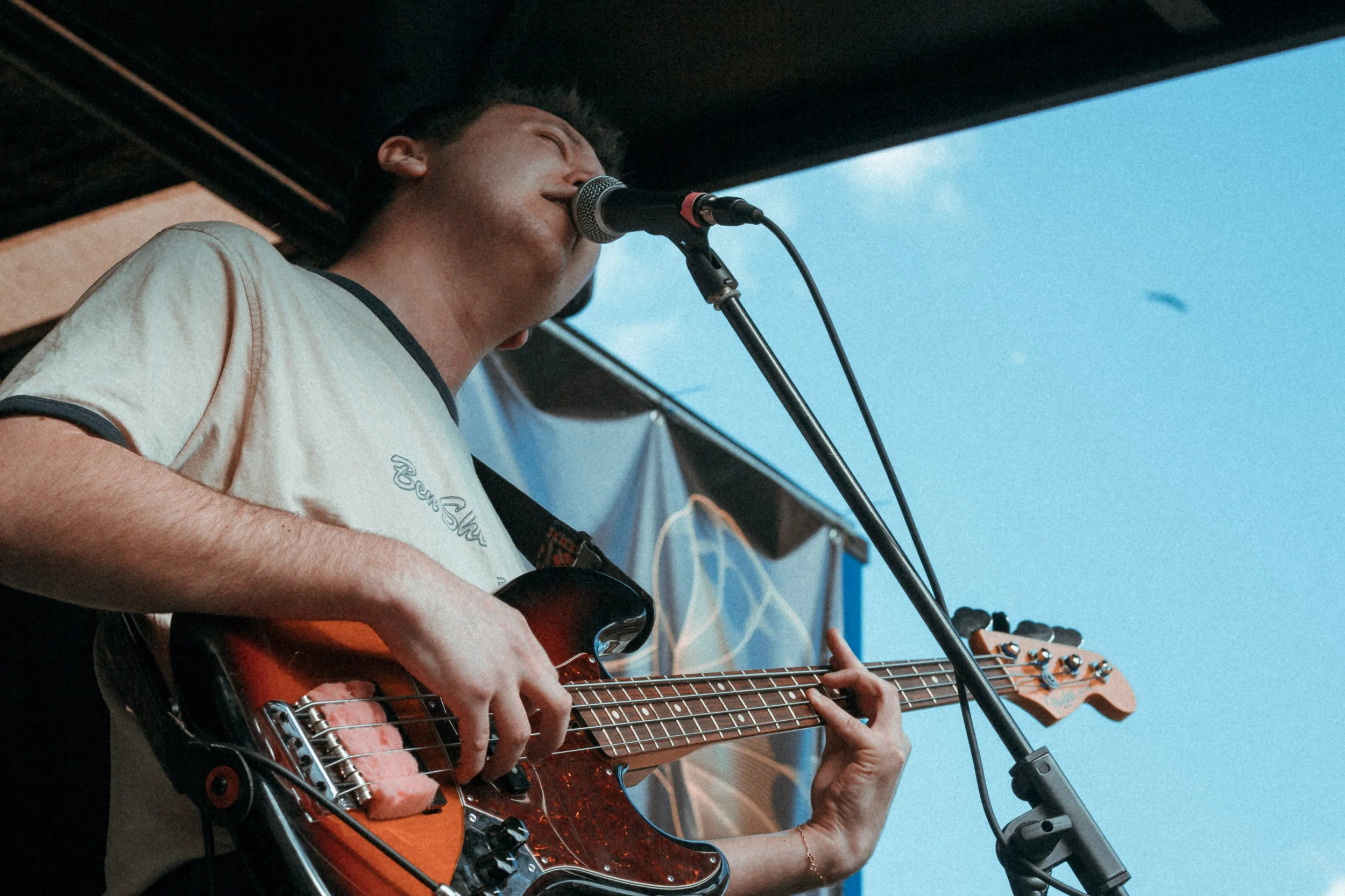 A young man performing live, singing into a microphone while playing an electric bass guitar outdoors.