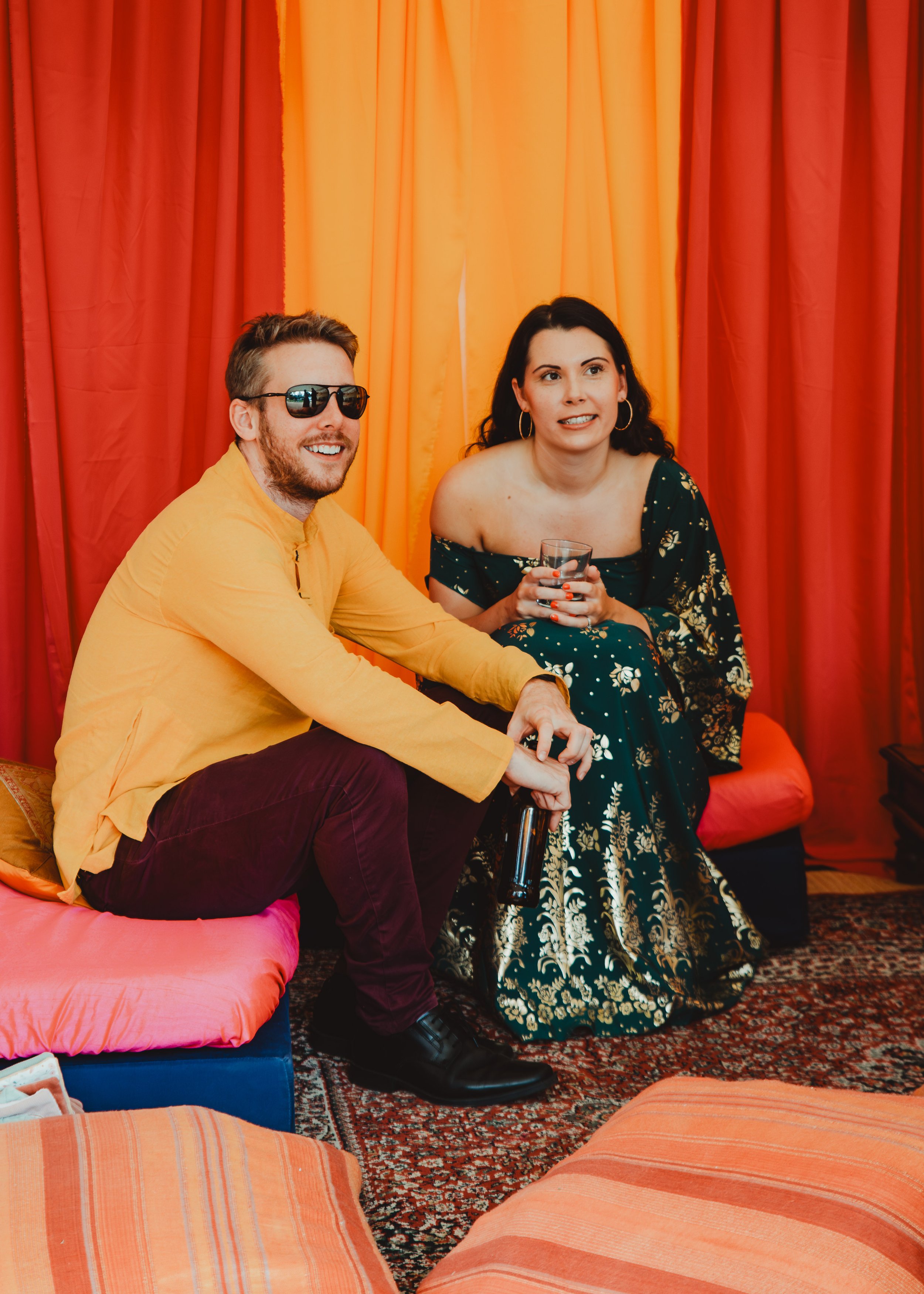 Two people sitting on colorful cushions in front of a vibrant red and yellow curtain, one man wearing sunglasses and a yellow shirt, smiling, and a woman holding a glass, wearing an off-the-shoulder floral dress, with a relaxed and cheerful demeanor.