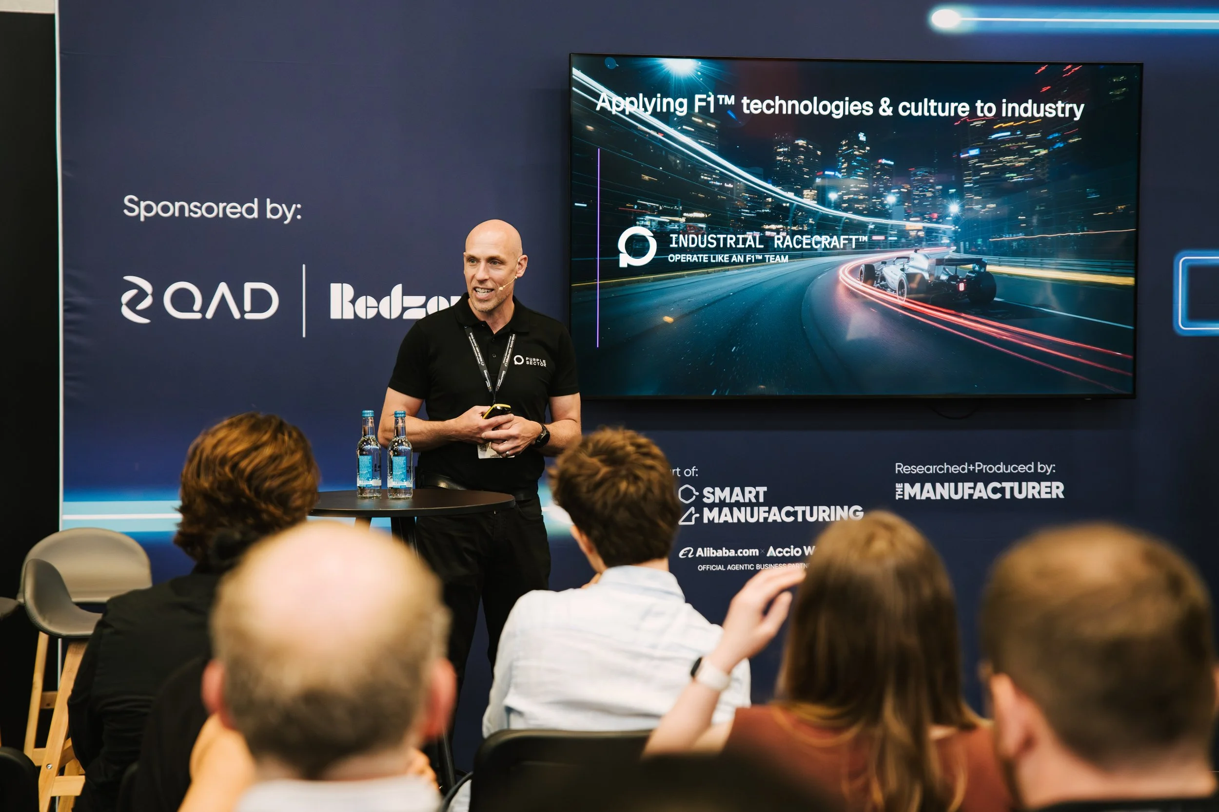 A man in a black shirt presenting at a conference, standing in front of a large screen displaying a graphic of a race car on a city street at night, with text about applying F1 technologies and culture to industry.