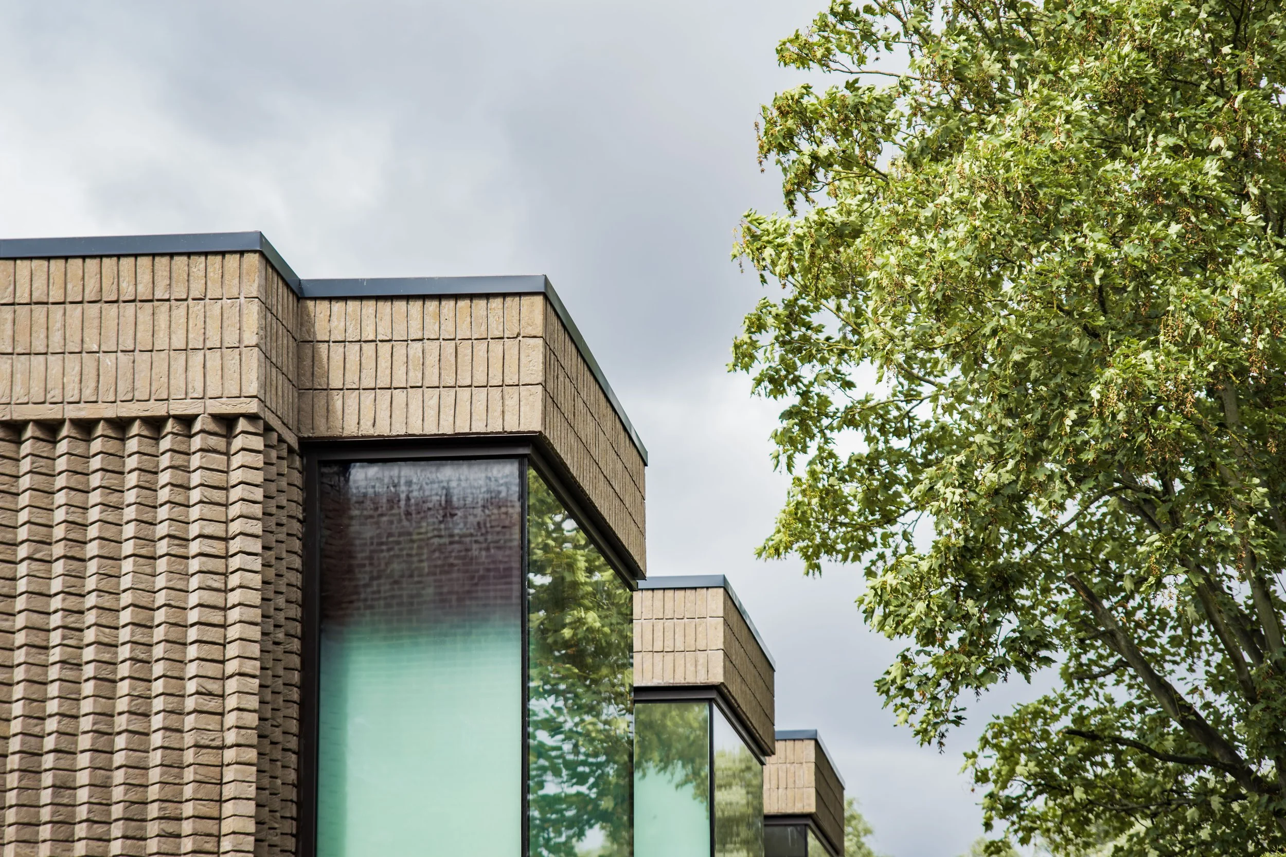 Close-up of a modern building corner with brick and glass window, partially obscured by green trees and an overcast sky.