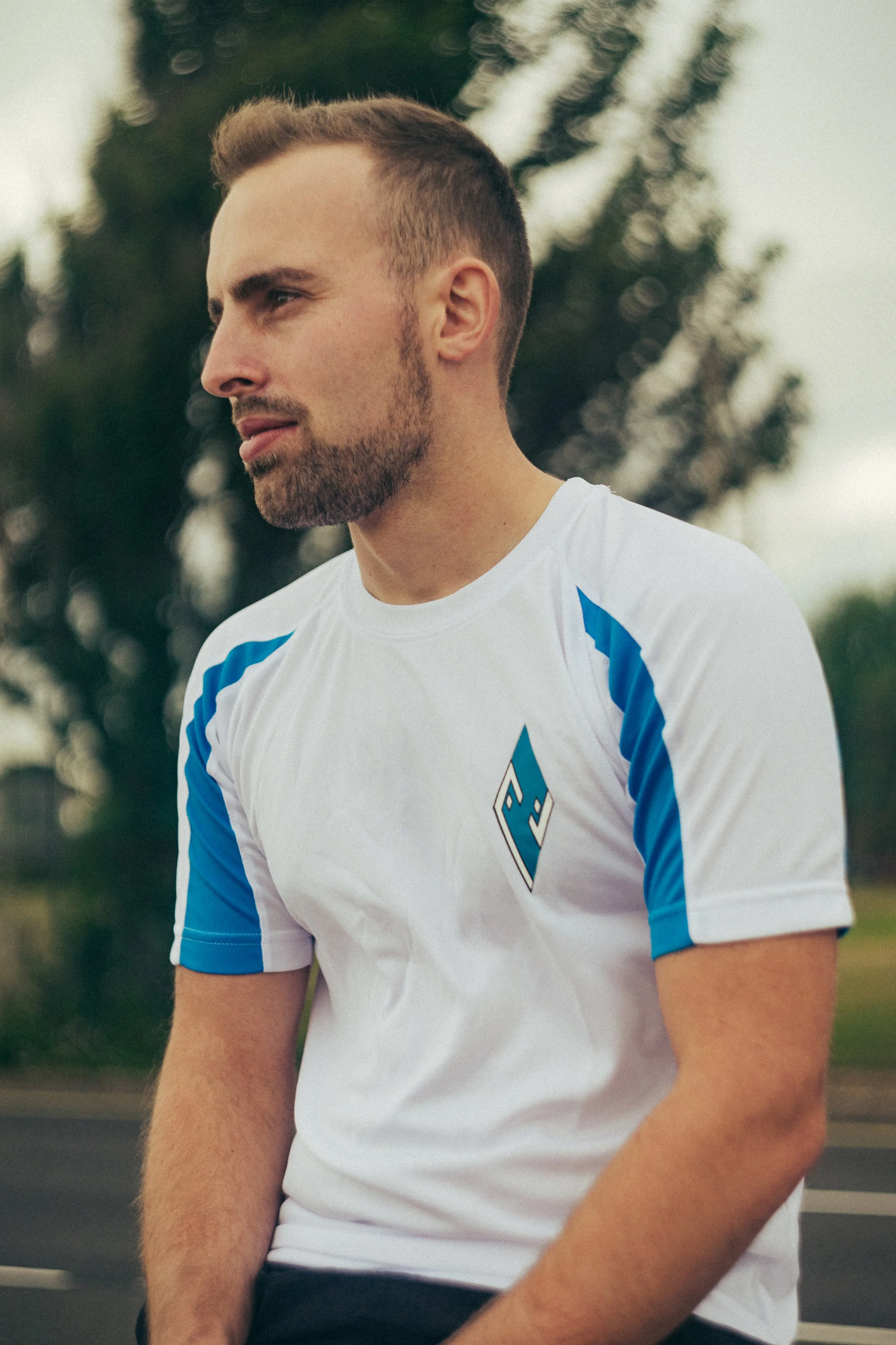 A young man with short, styled hair and a beard, wearing a white sports jersey with blue accents, looking to the side outdoors with trees in the background.