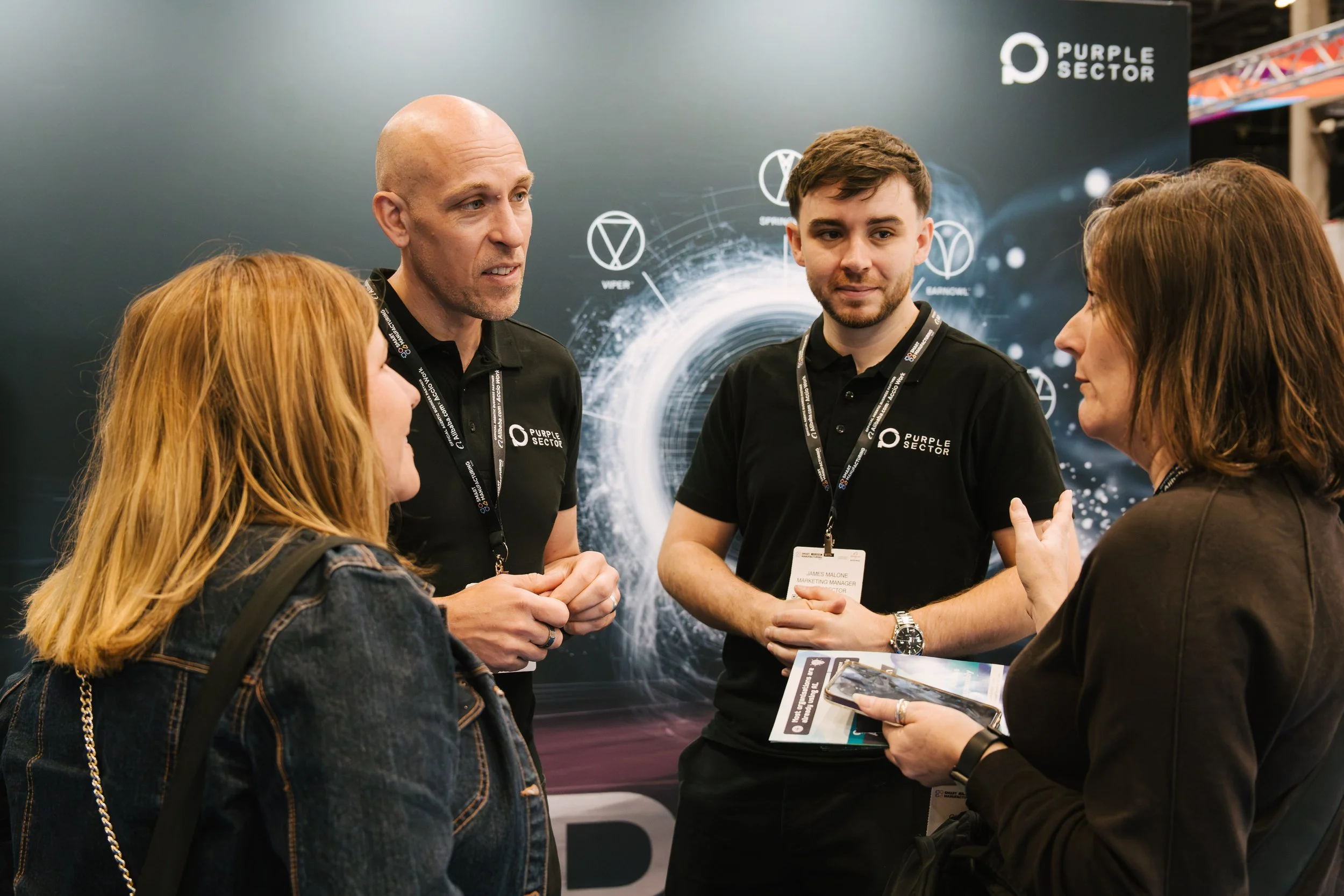 A group of five people, three women and two men, engaged in conversation at a trade show or conference. The background shows a dark blue booth with logos and futuristic graphics. The men are wearing black shirts with the "Purple Sector" logo, and some individuals are holding brochures or smartphones.