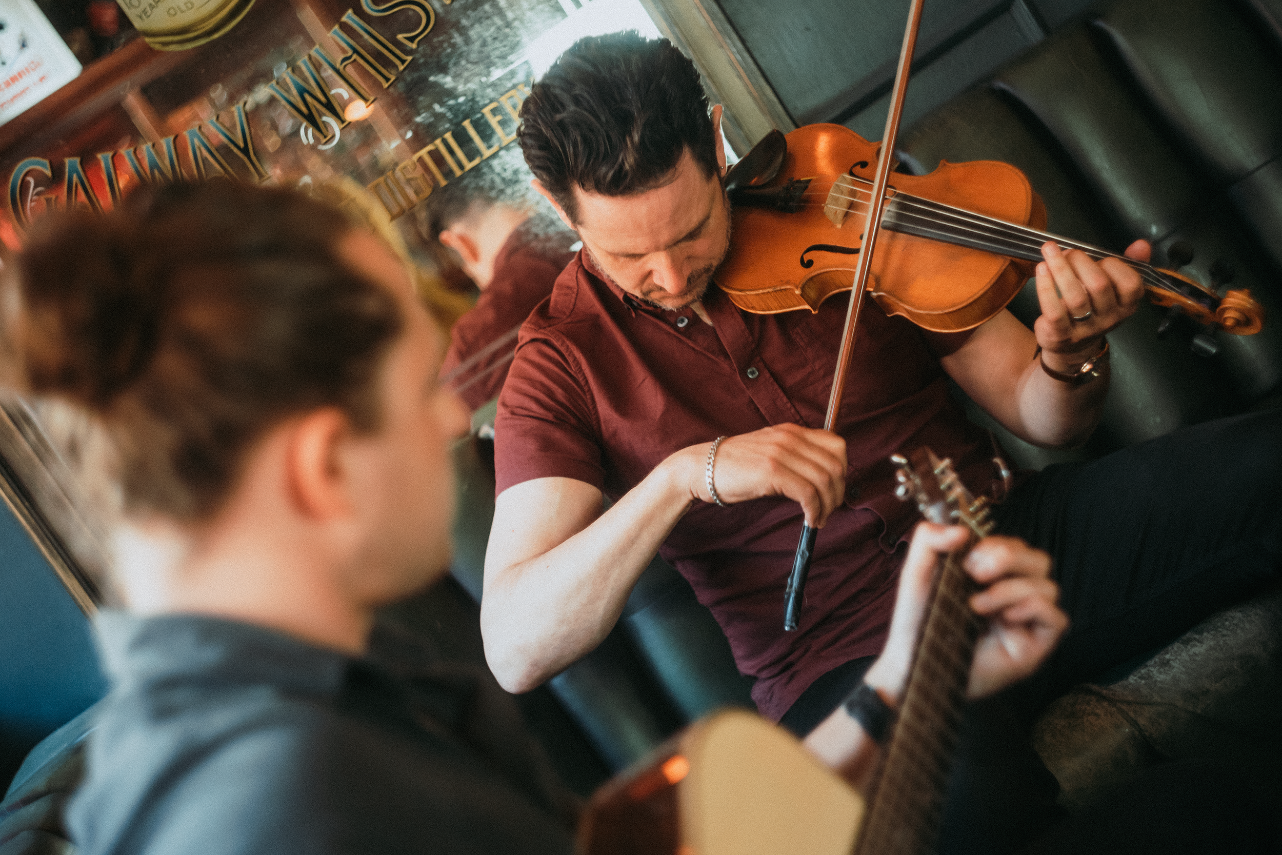 A man with dark hair playing a violin with a bow, sitting on a bus or train next to a woman, with a window showing a sign and trees outside. The man is wearing a maroon shirt and a bracelet.