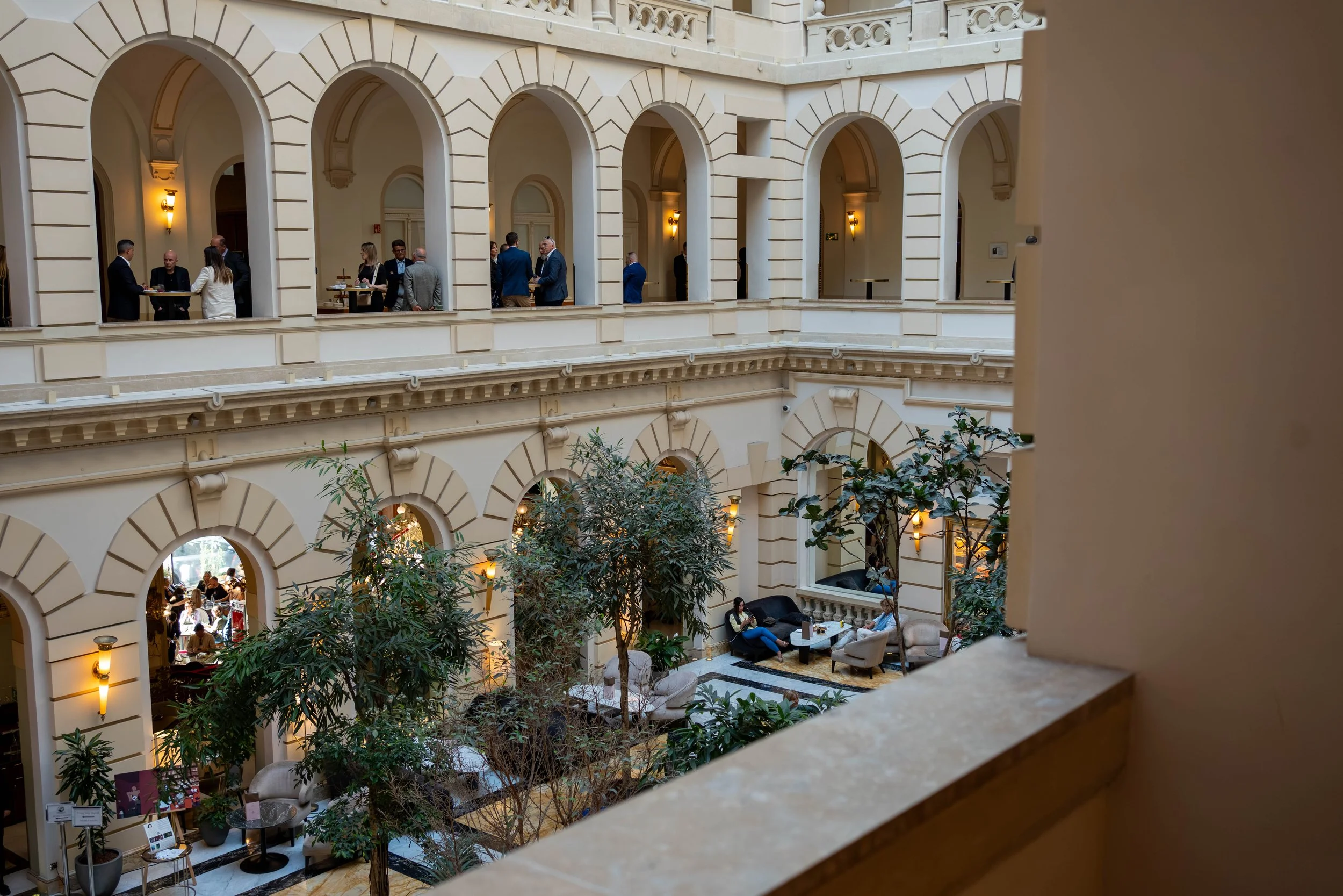 View of an elegant indoor atrium with multiple floors featuring cream-colored walls, large arched windows, ornate architectural details, and lush greenery; several groups of people are engaged in conversation and sitting in the lounge area.