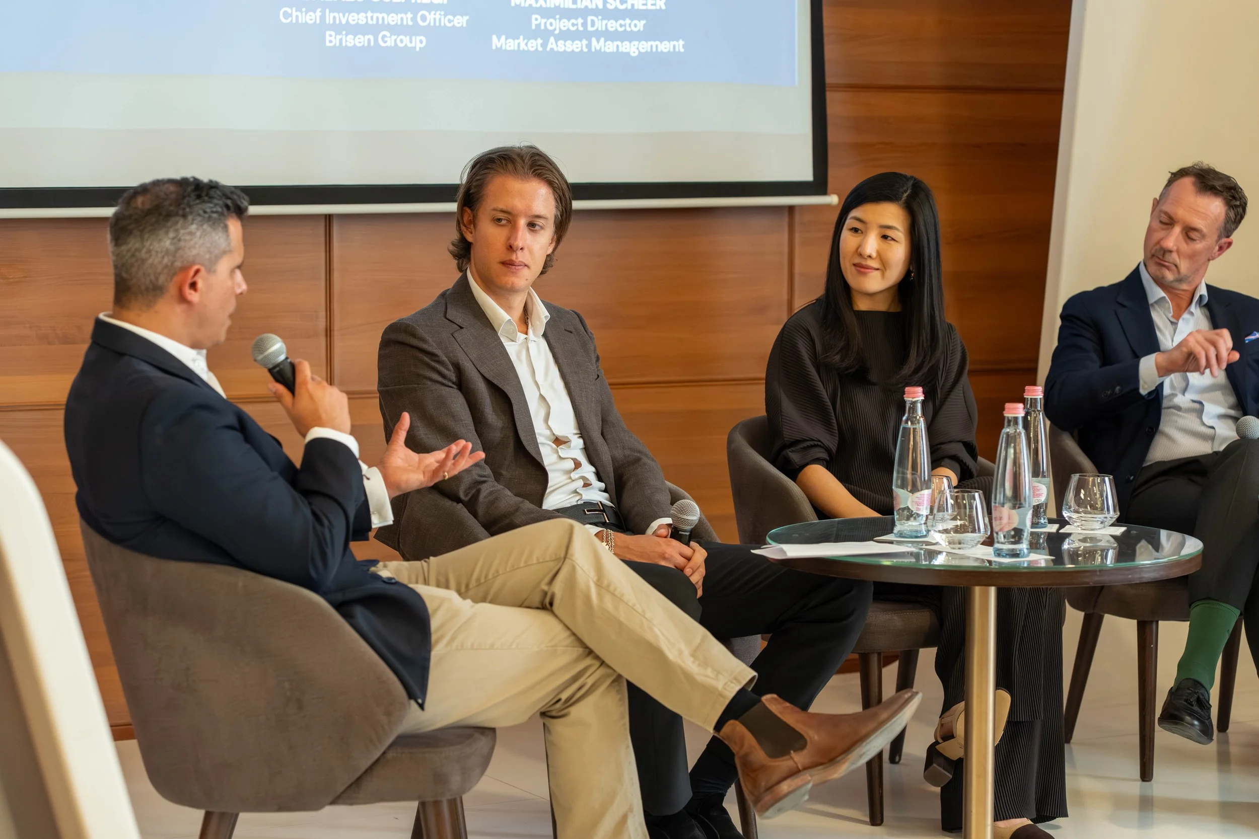 Four people seated at a panel discussion or meeting, with microphones, water bottles, and glasses on the table. A presentation slide is partially visible in the background.