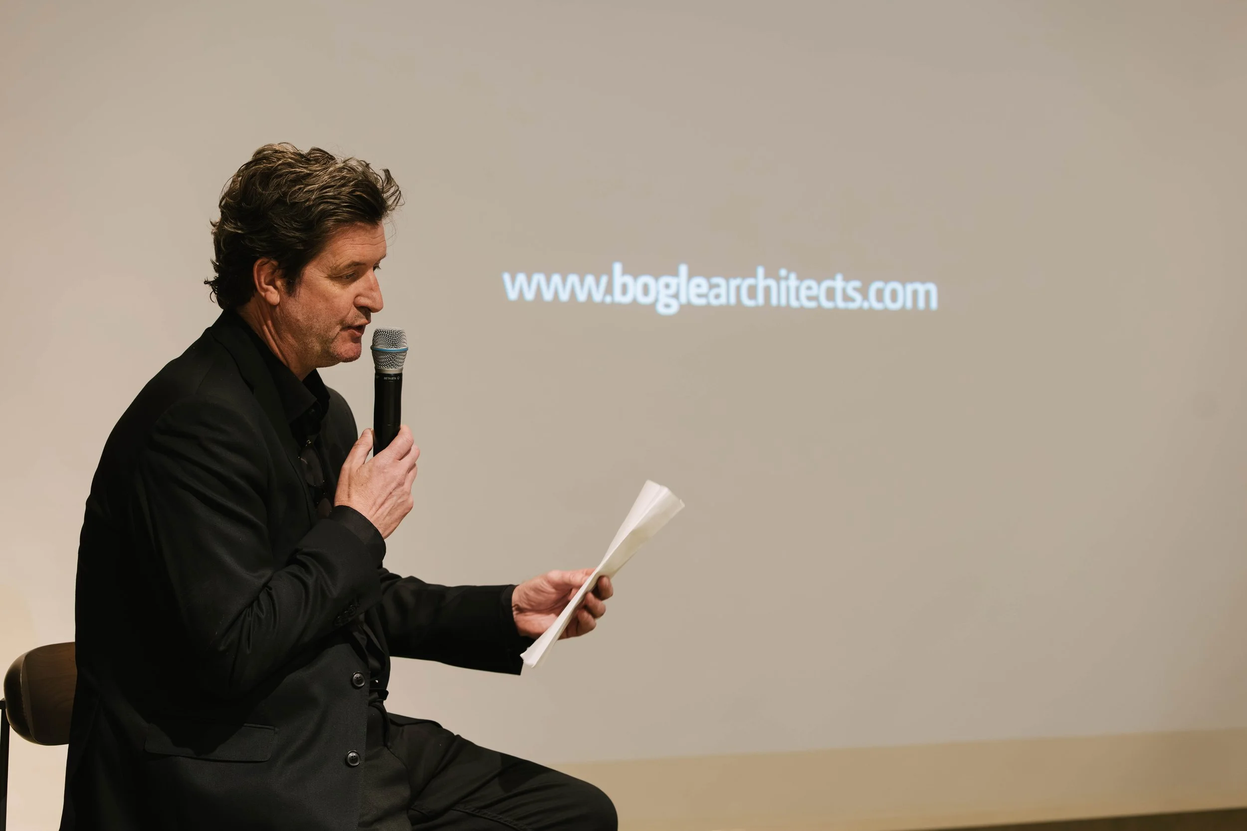 A man in a black suit sitting on a chair, holding a microphone in one hand and reading from a paper in the other, with a presentation slide displaying a website URL in the background.