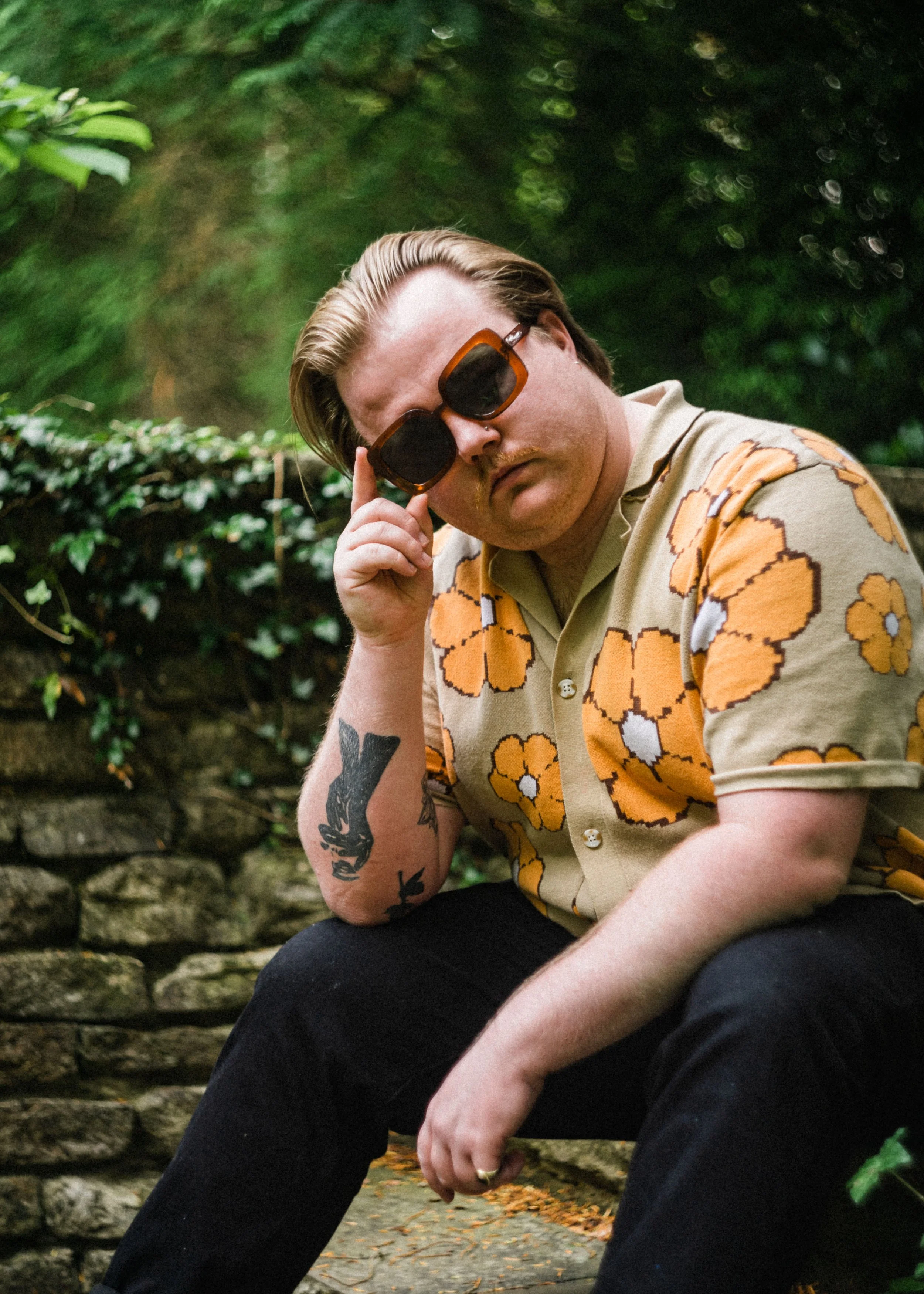 A man with blonde hair and a mustache, wearing large tortoise shell sunglasses and a beige shirt with orange and white floral pattern, sits outdoors on a stone ledge in a lush green setting.
