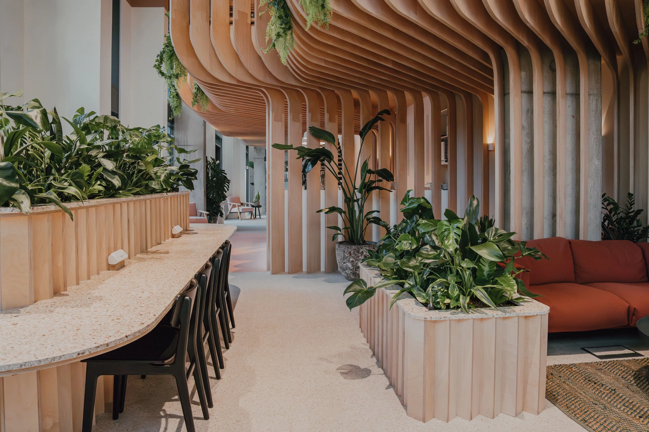 Interior of a modern, stylish space with a curved wooden ceiling, lush green plants, a long counter, and seating area with chairs and a red sofa.