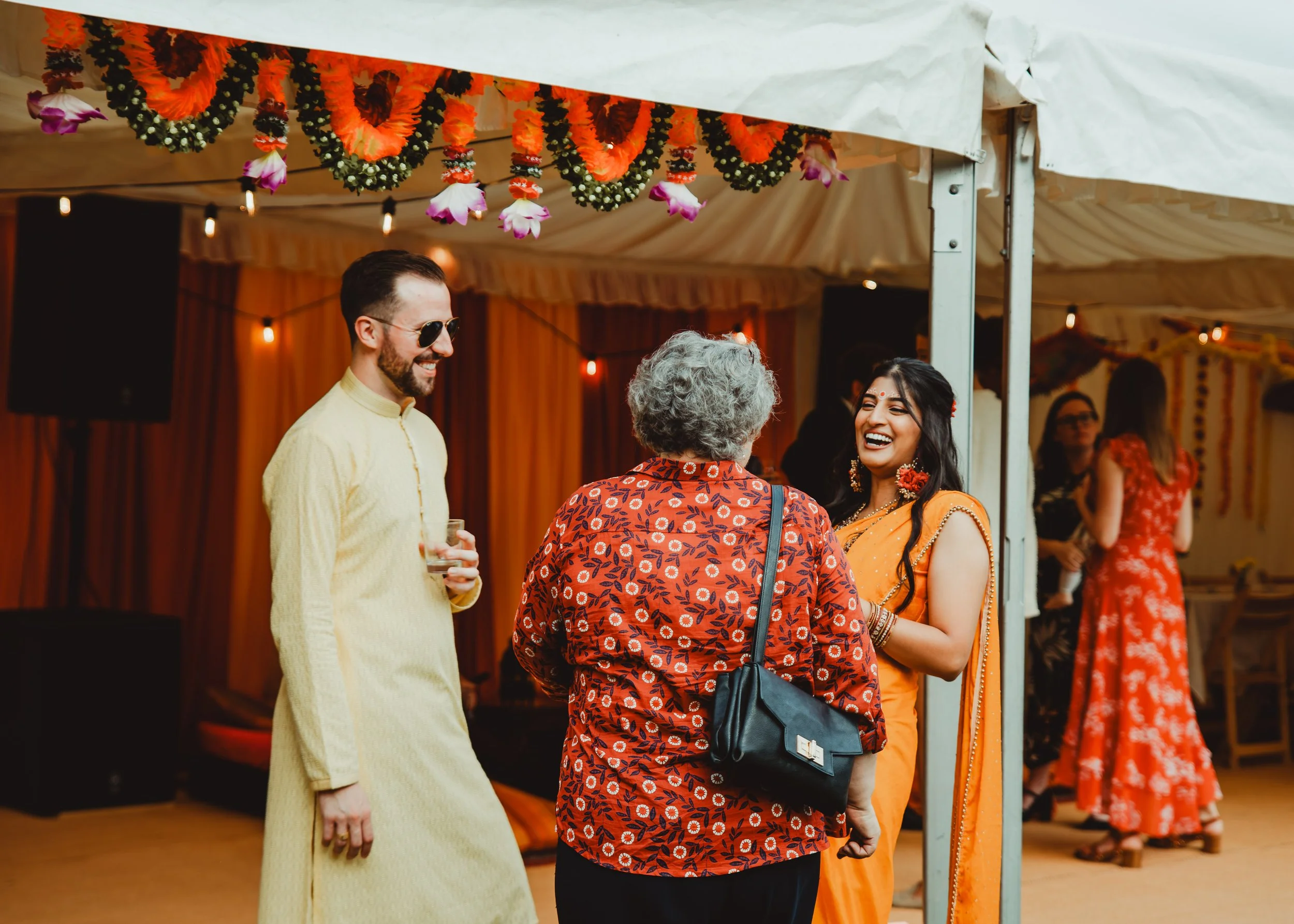 Two women and a man at a celebration or cultural event, decorated with vibrant orange and pink flowers and string lights, engaging in conversation inside a decorated tent.