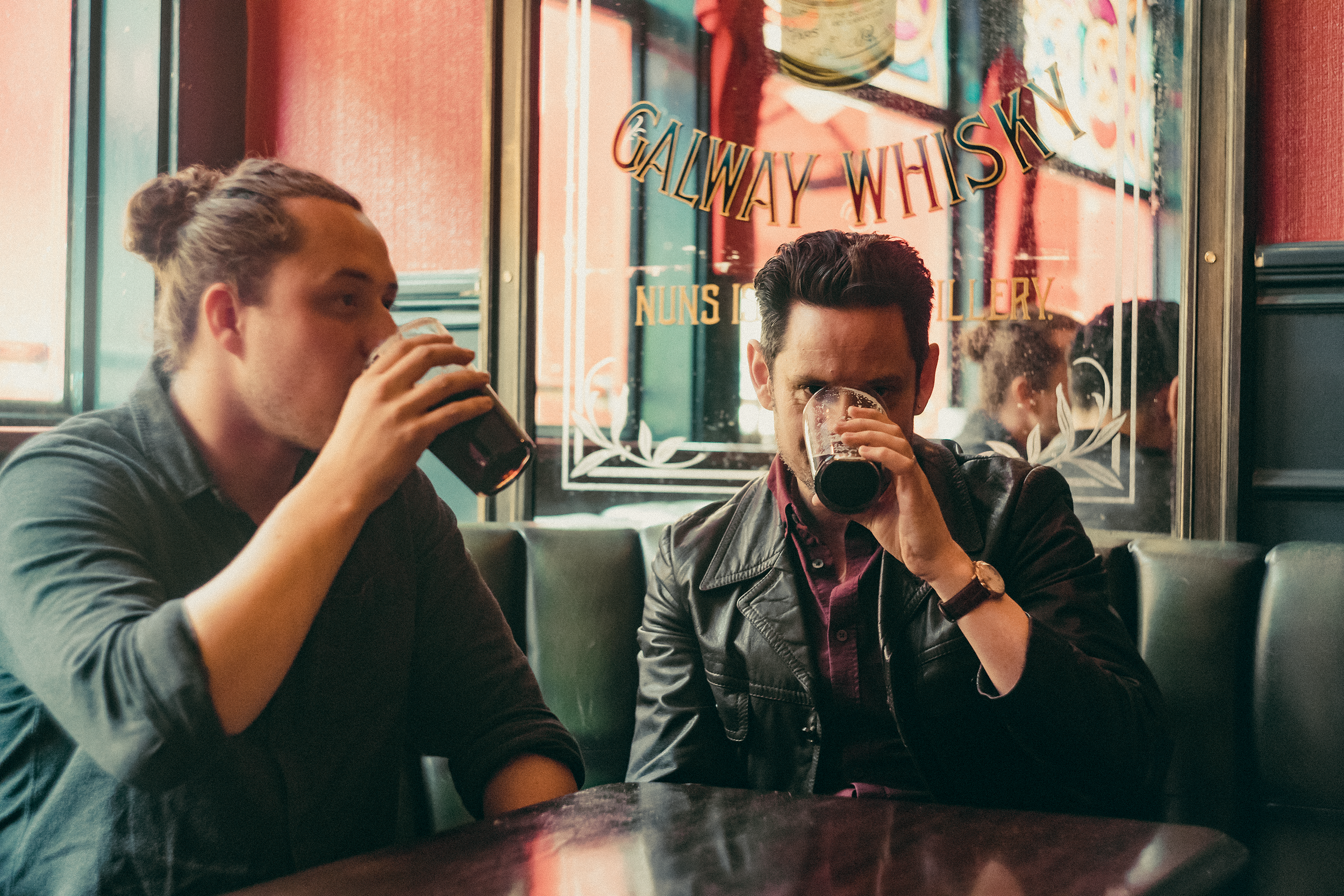 Two men sitting in a diner, drinking dark beverages, with a mirror behind them reflecting the scene and decorated with the words 'GALLERY WISBY' in a colorful, stylized font.