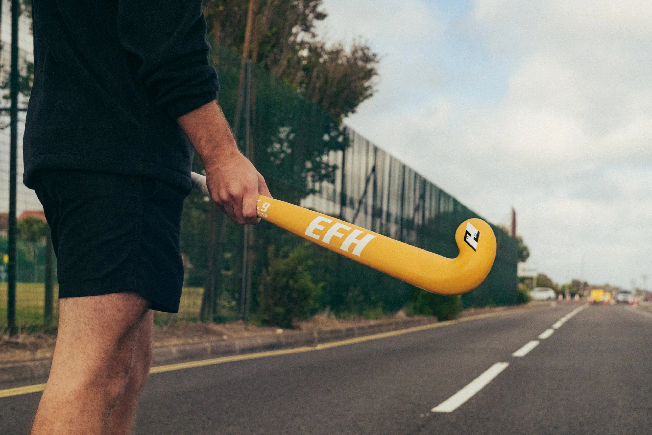 A person holding a yellow field hockey stick walking along a road with trees and a large building in the background.