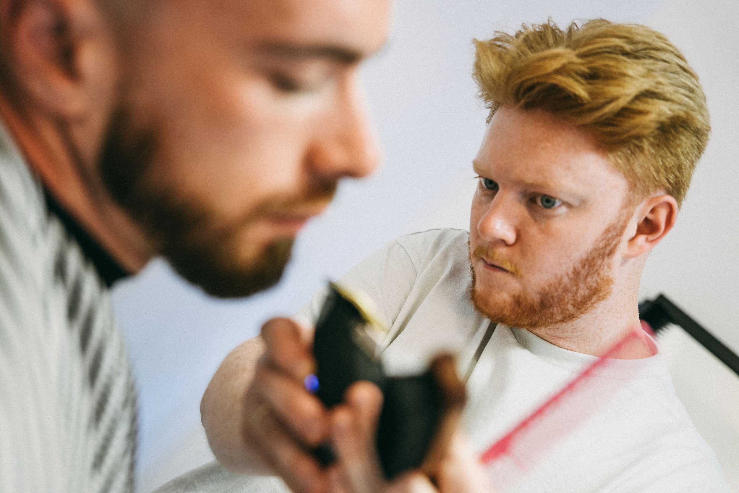 Two men with red hair and beards are looking at a phone together. One is slightly out of focus in the foreground, and the other is in focus, looking at the phone screen. The background is plain and white.