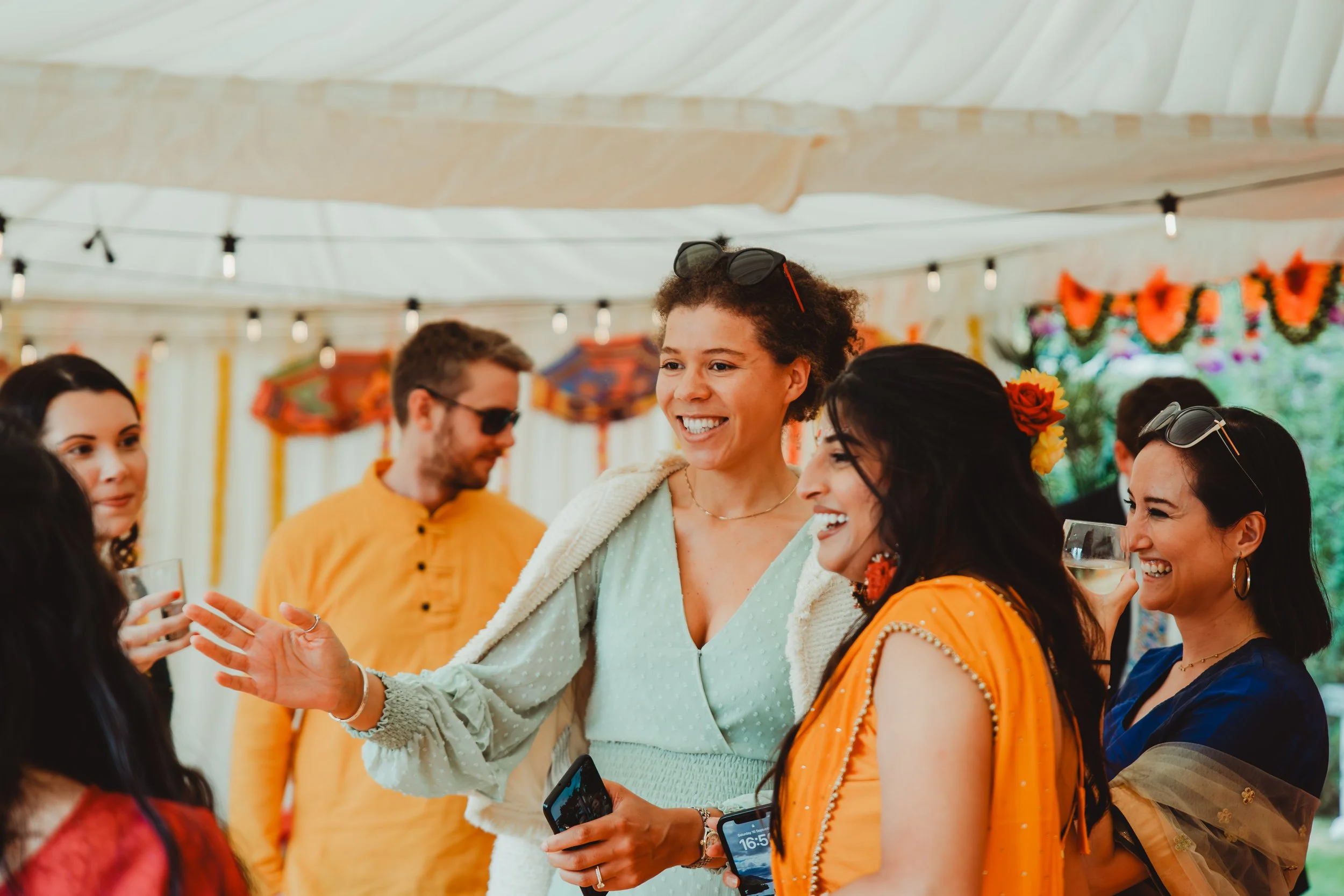 People celebrating at a colorful outdoor event, with women wearing bright traditional dresses and floral accessories, smiling and engaging with each other.
