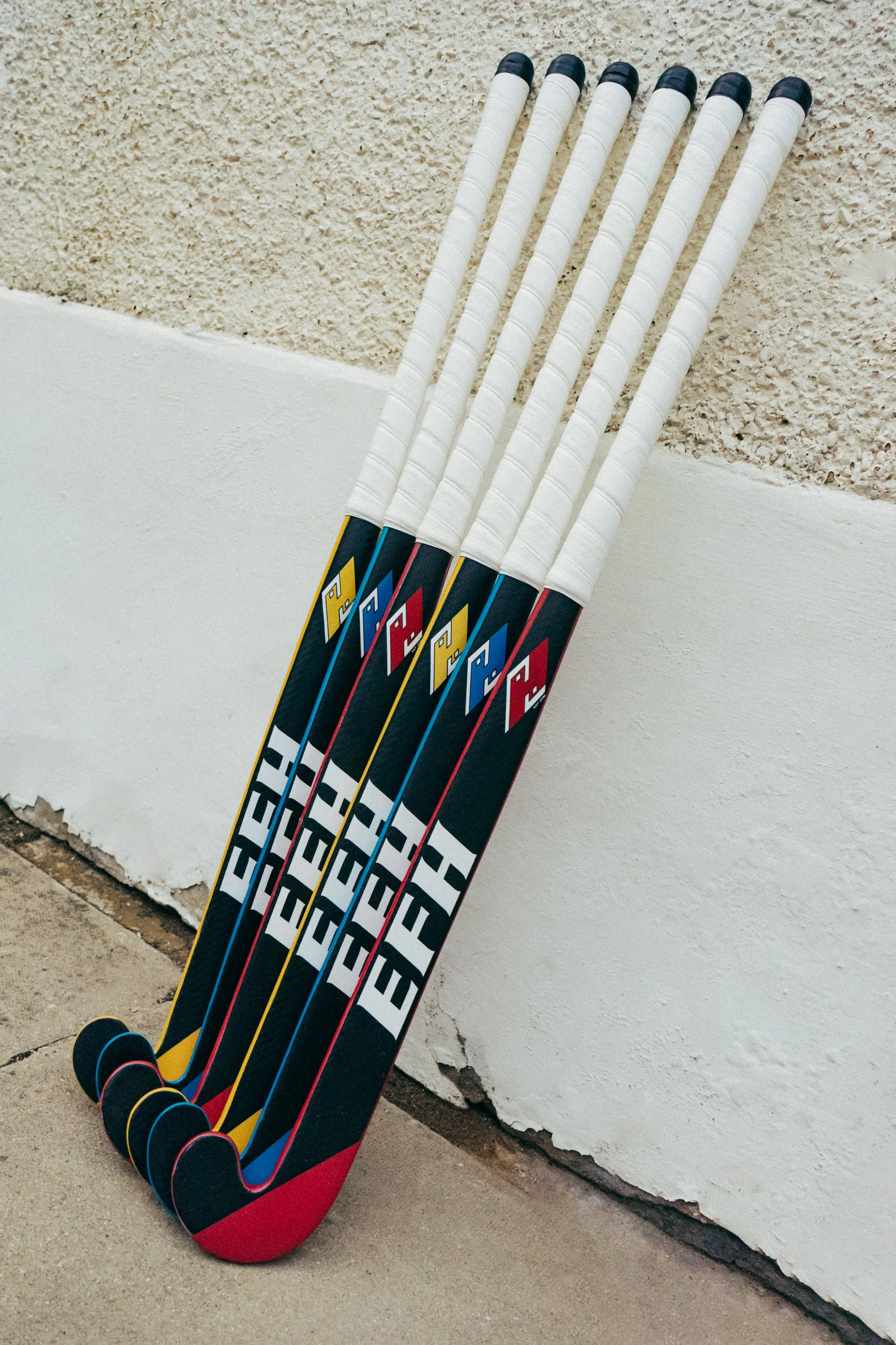 Five colorful hockey sticks leaning against a beige wall with a white ledge at the bottom.